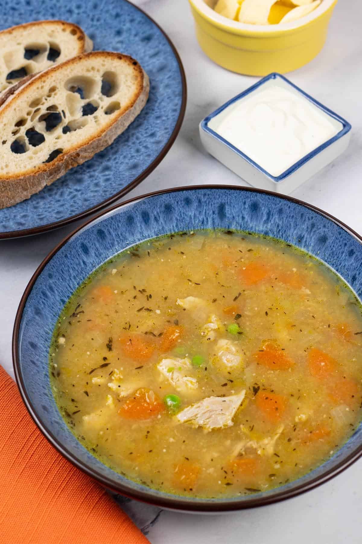 A portion of slow cooker roast chicken soup in a blue patterned bowl with a plate of sliced Portuguese water bread and dish of garlic and lemon yogurt on the side.