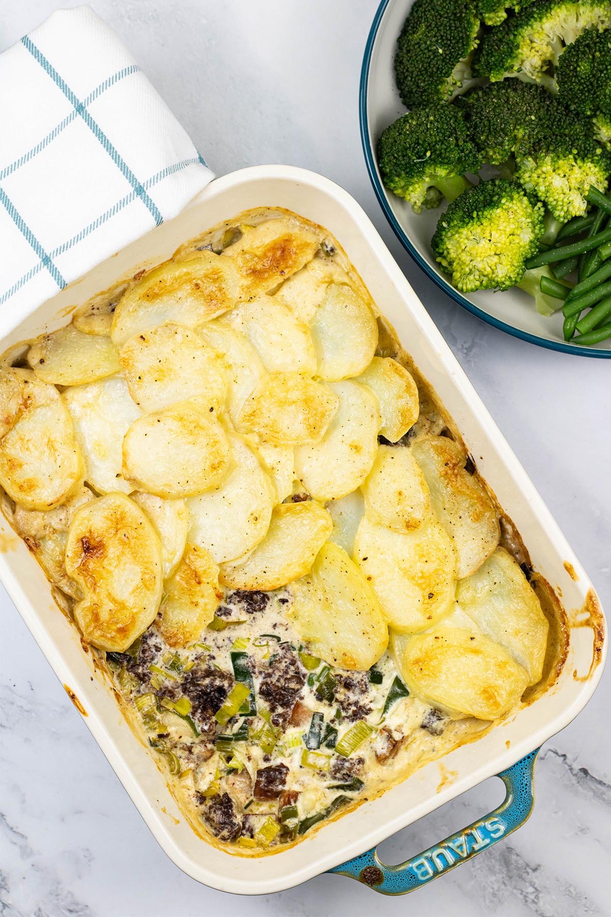 Smoked haddock, black pudding and leek gratin in a rectangle Staub dish with a bowl of broccoli and green beans on the side.