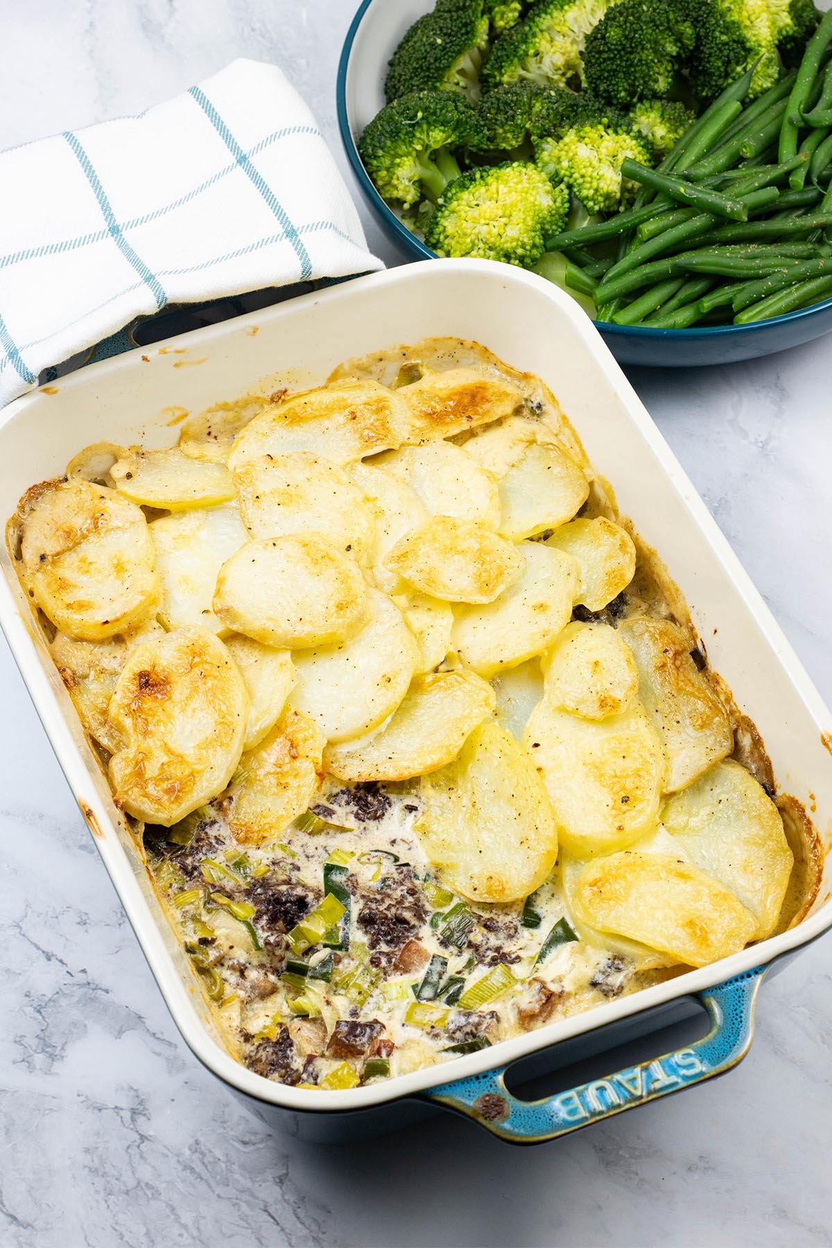 Smoked haddock, black pudding and leek gratin in a rectangle Staub dish with a bowl of broccoli and green beans on the side.