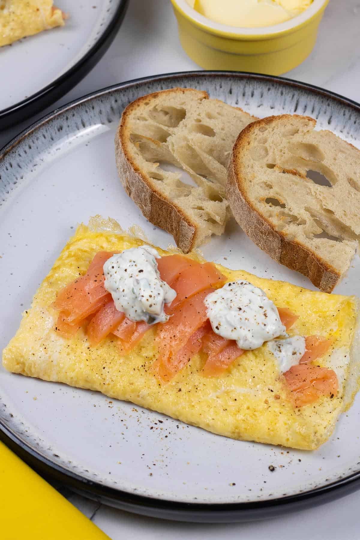 A portion of smoked salmon and lemon herb cream cheese omelette on a black and white patterned dinner plate with a slice of toasted Portuguese water bread.