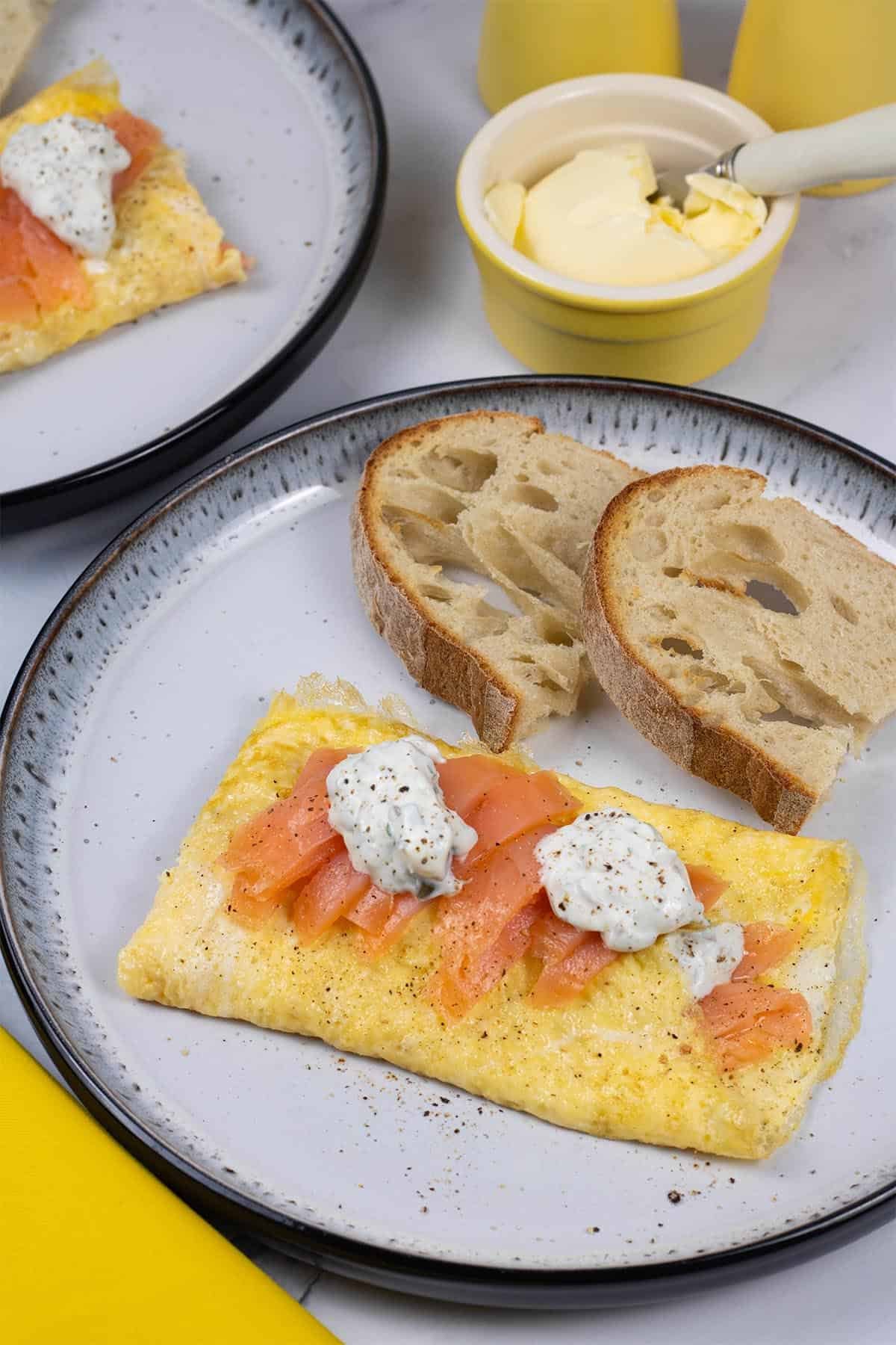 Two portions of smoked salmon and lemon herb cream cheese omelette on black and white patterned dinner plates with a slice of toasted Portuguese water bread.