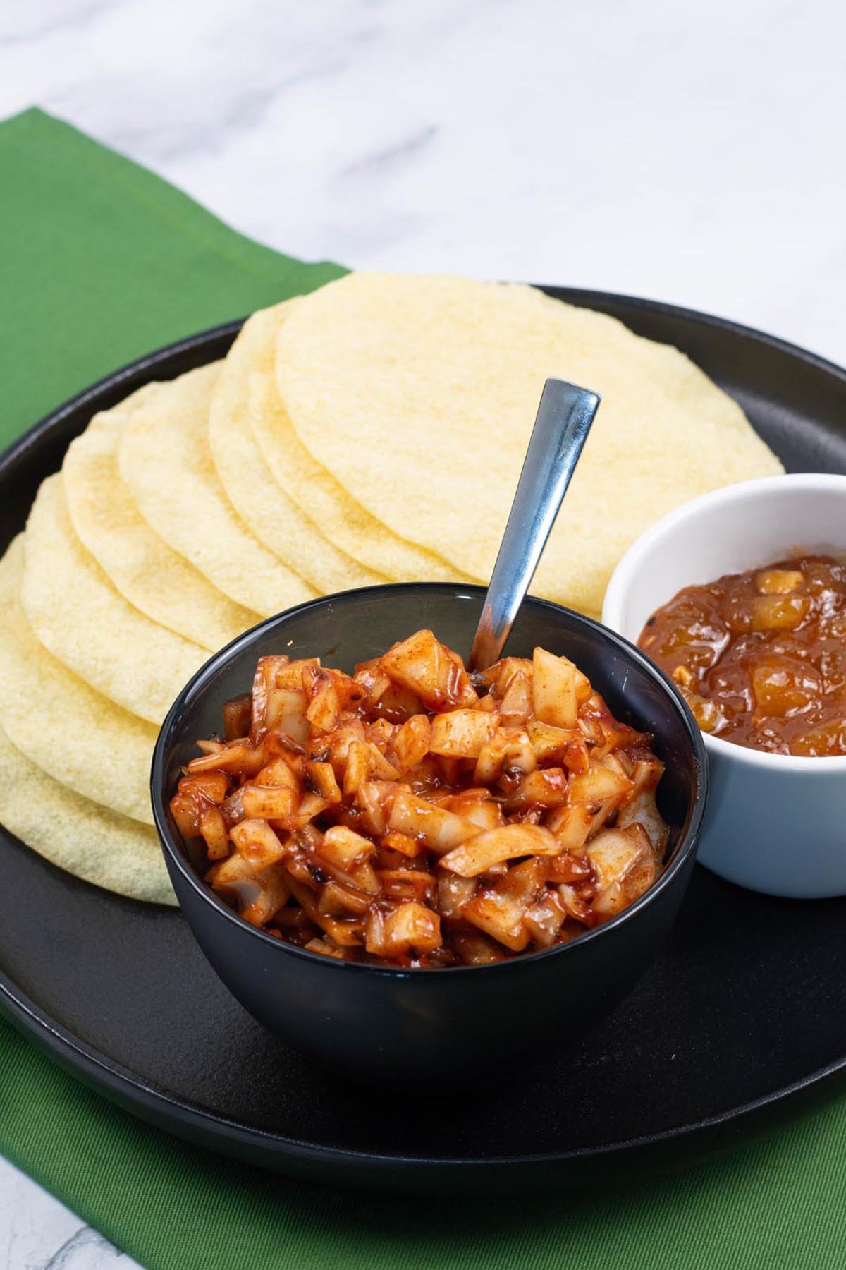 Spicy onions in a black serving bowl sitting on a black dinner plate with poppadoms and a dish of mango chutney.