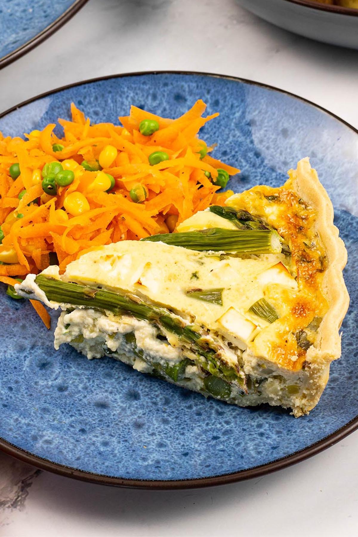 A slice of spring vegetable and feta quiche on a blue patterned side plate with some warm carrot salad on the side.