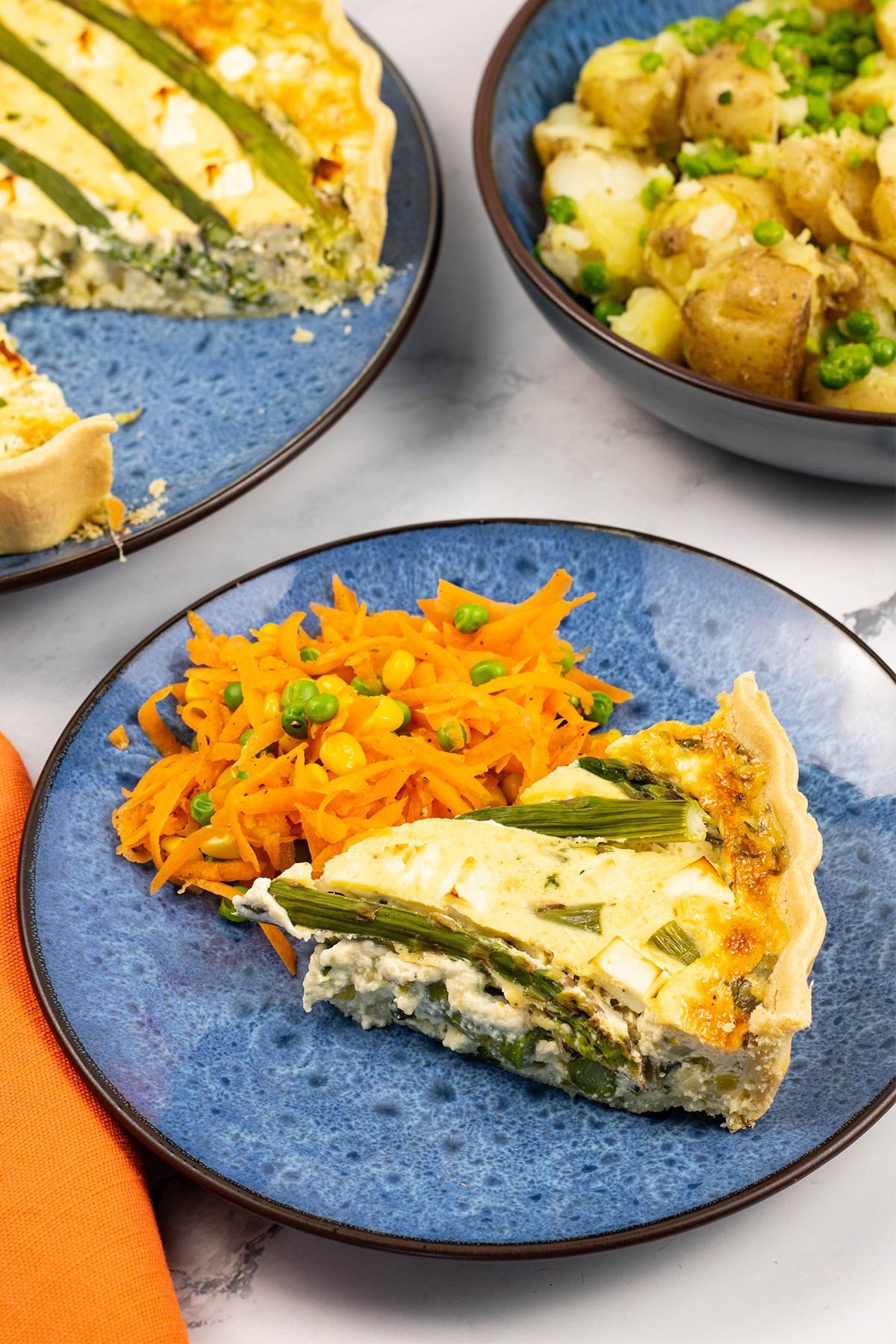 A slice of spring vegetable and feta quiche on a blue patterned side plate with some warm carrot salad on the side.