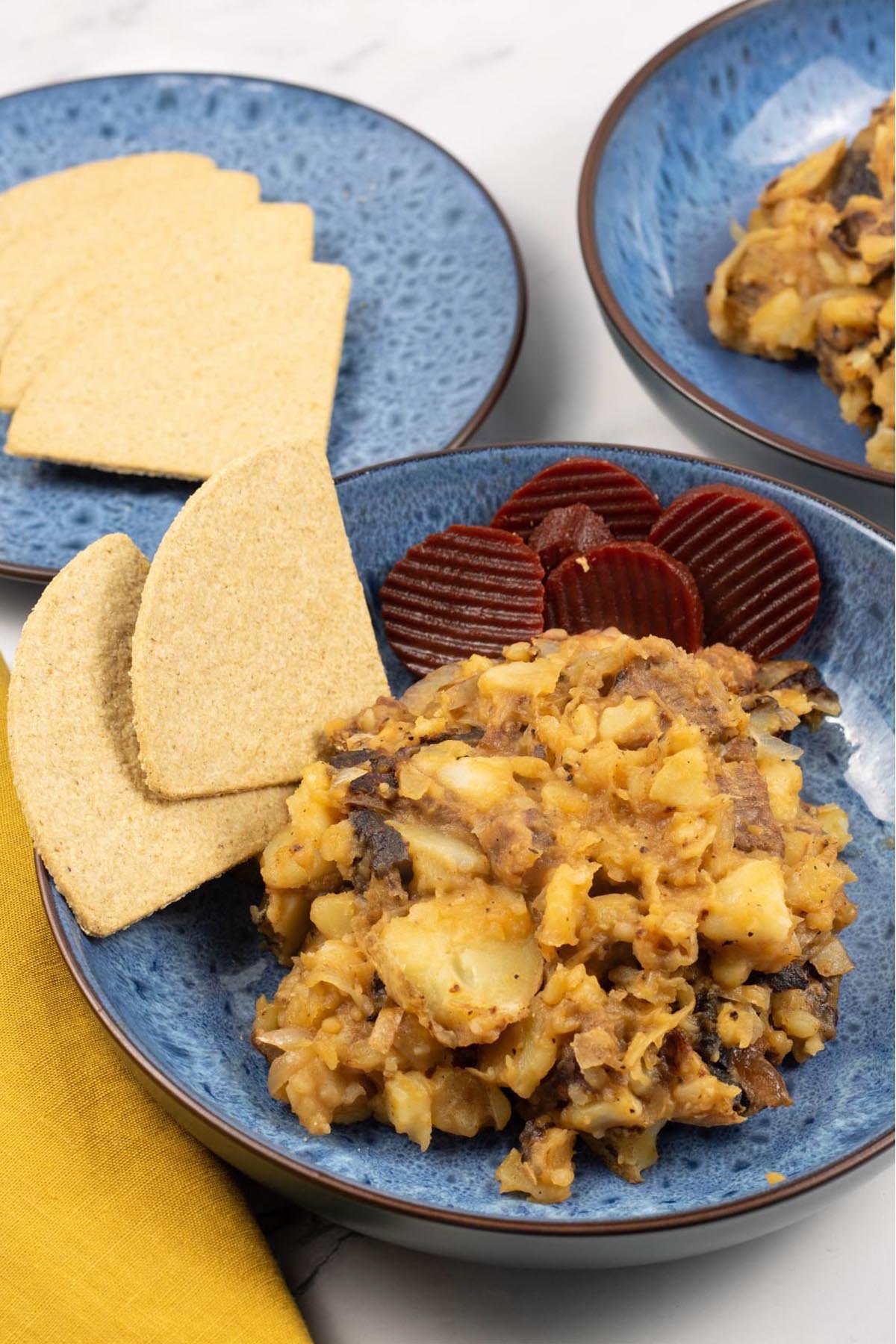 Two portions of stovies in blue patterned bowls, garnished with sliced beetroot and oatcakes.