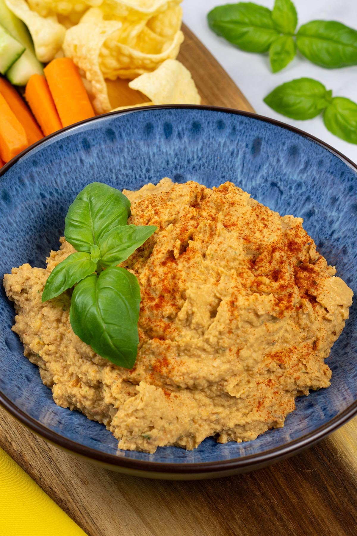 Sun-dried tomato and basil houmous in a blue patterned serving bowl sitting on a board with carrot and cucumber sticks and lentil crisps.