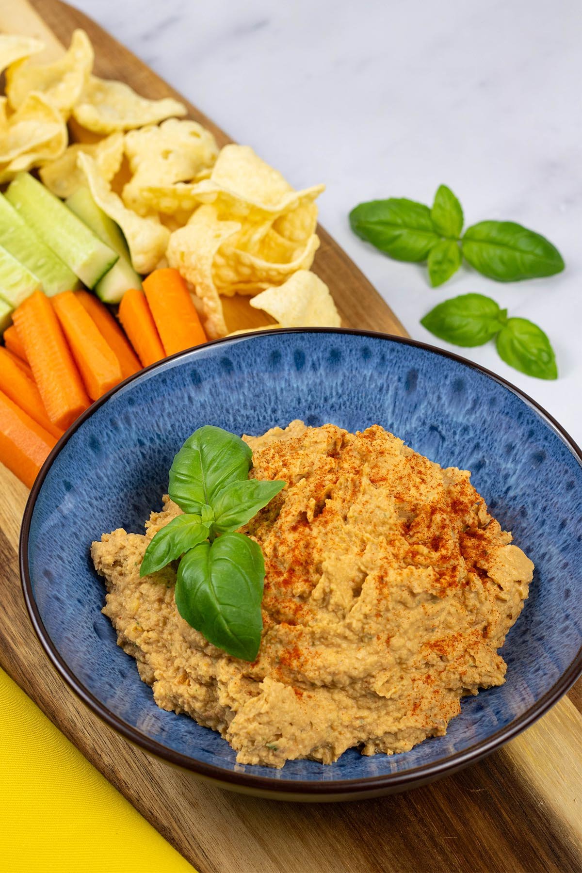 Sun-dried tomato and basil houmous in a blue patterned serving bowl sitting on a board with carrot and cucumber sticks and lentil crisps.