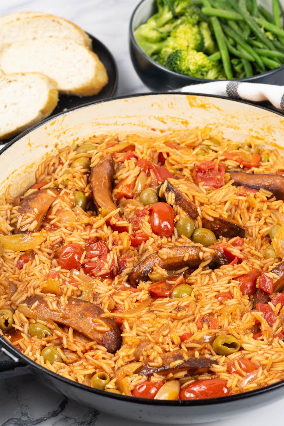 Tomato and chorizo rice in a large shallow round casserole with a bowl of broccoli and green beans, and plate of sliced crusty baguette in the background.
