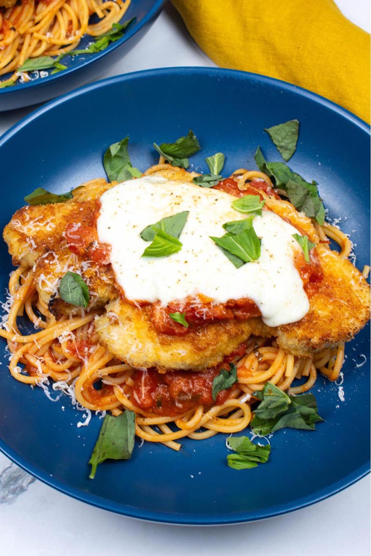 Close up of turkey parmigiana with spaghetti in a blue bowl with a mustard napkin on the side.