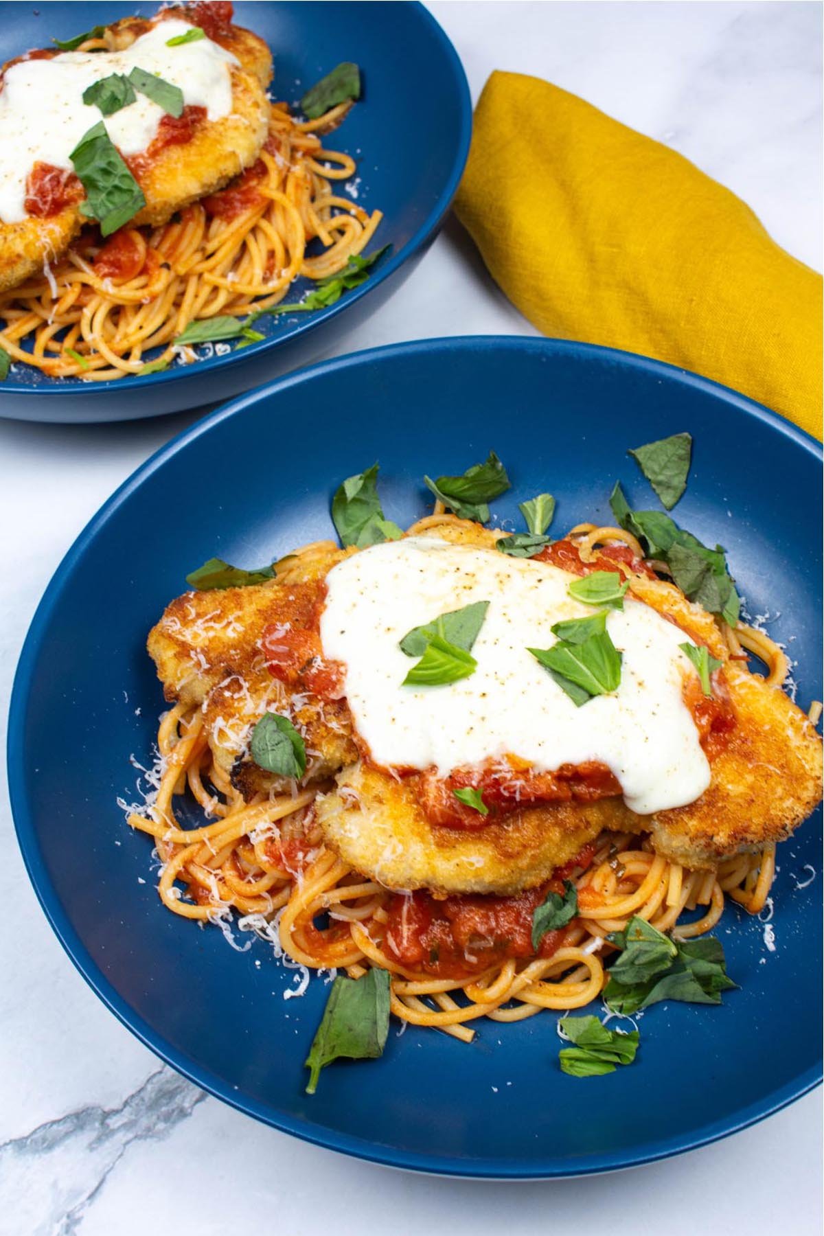 2 bowls of turkey parmigiana with spaghetti in blue bowls with a mustard napkin on the side.