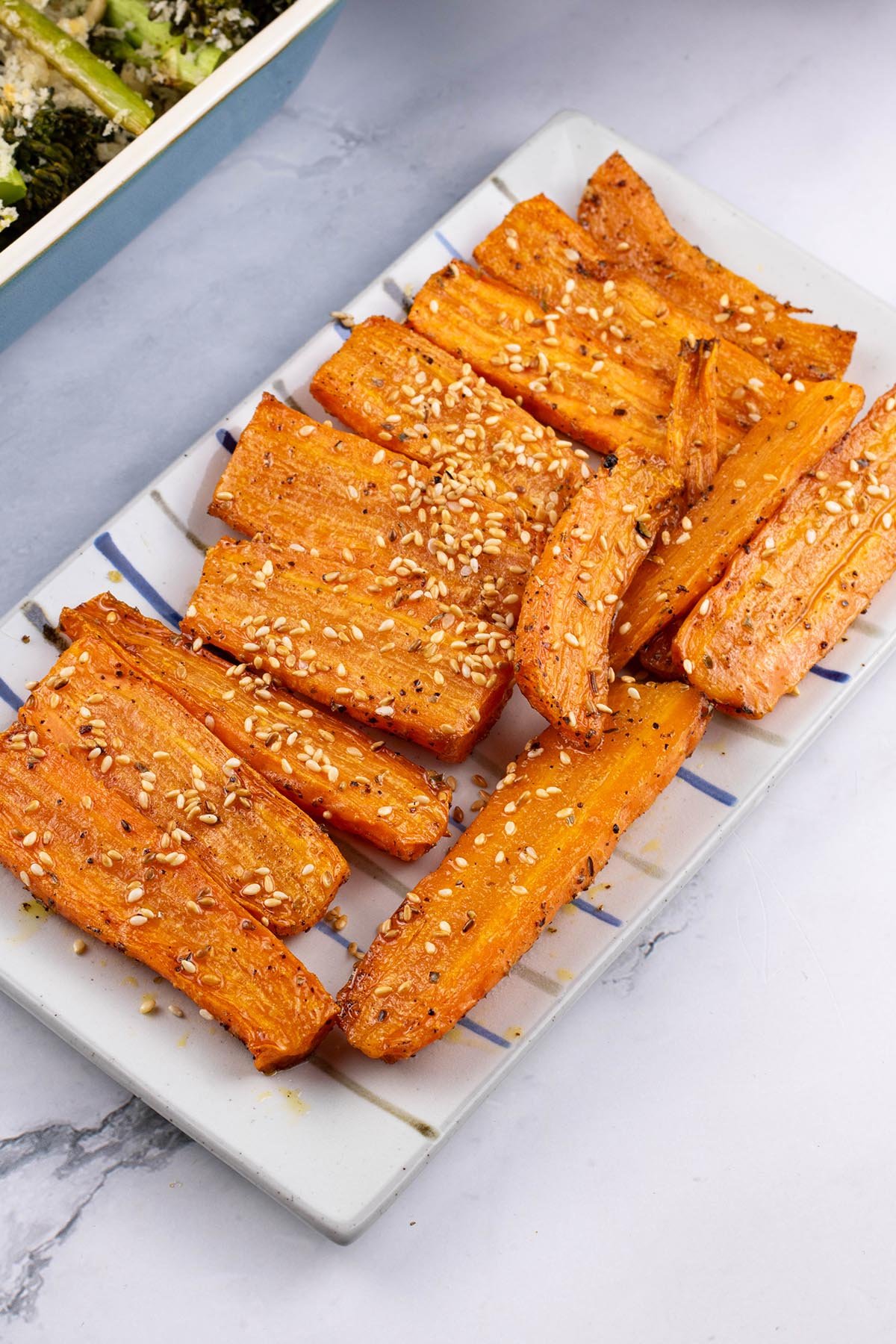 Za'atar roasted carrots on a rectangle striped platter with a dish of tenderstem broccoli on the side.