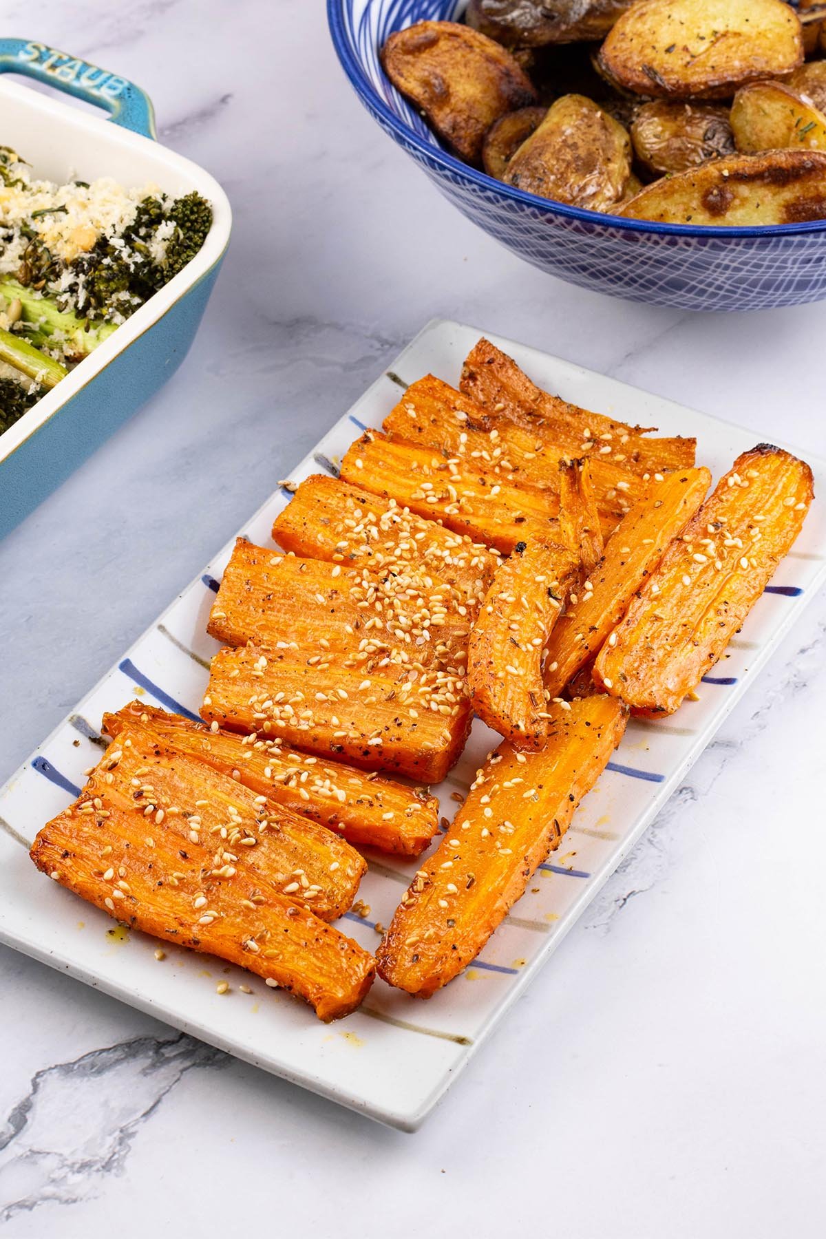 Za'atar roasted carrots on a rectangle striped platter with a dish of tenderstem broccoli and bowl of sautéed potatoes on the side.
