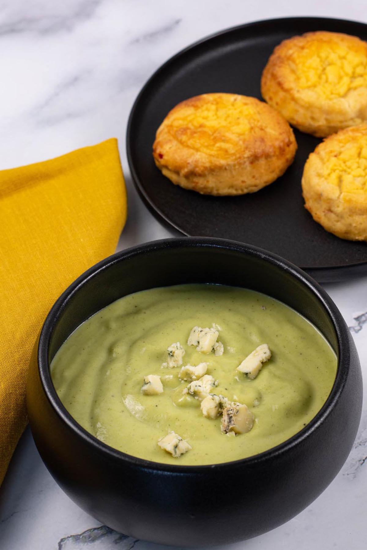A portion of broccoli and stilton soup garnished with crumbled stilton in a black bowl, made in the Thermomix, with a plate of chilli cheese scones on the side.