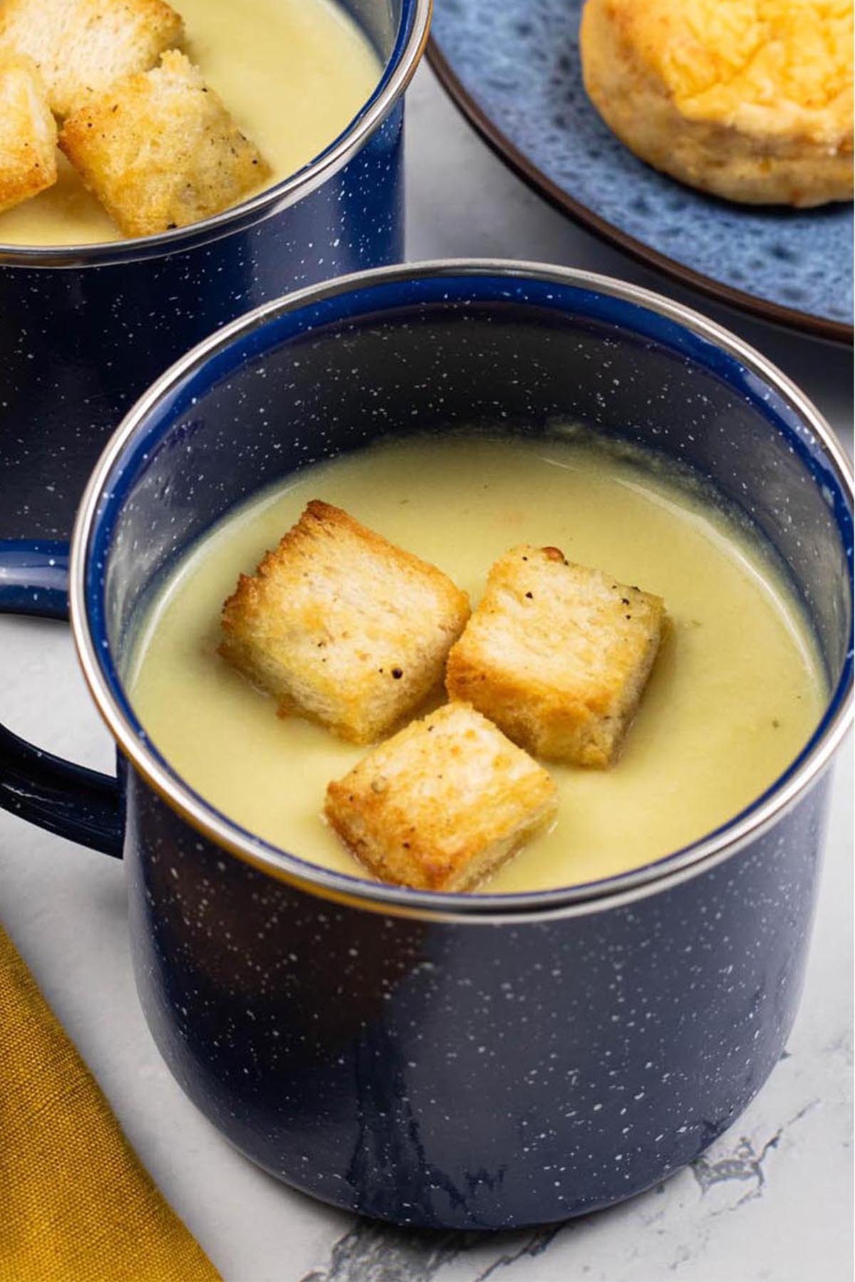 Two portions of cauliflower cheese soup in blue enamel mugs with a plate of chilli cheese scones on the side.