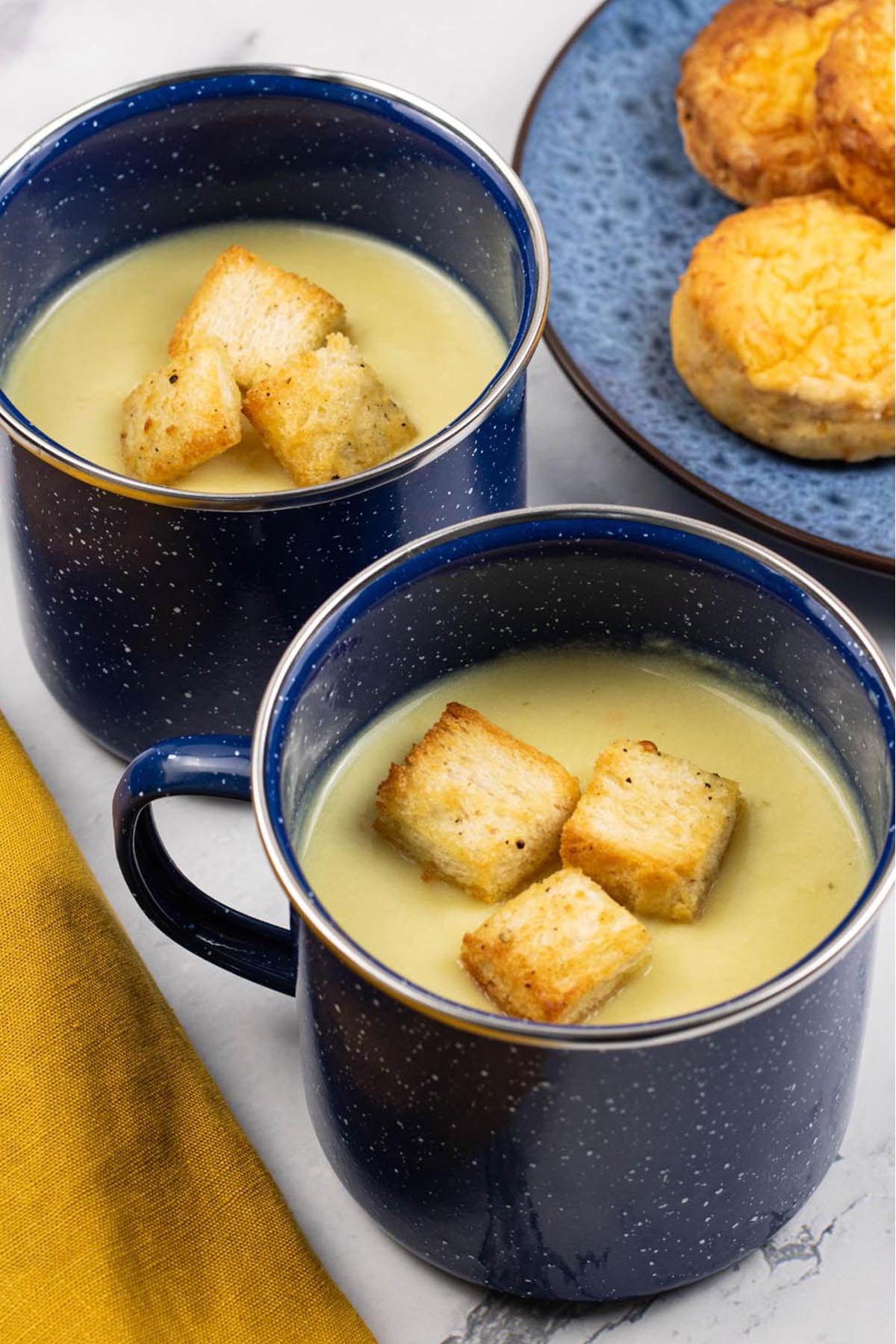 Two portions of cauliflower cheese soup in blue enamel mugs with a plate of chilli cheese scones on the side.