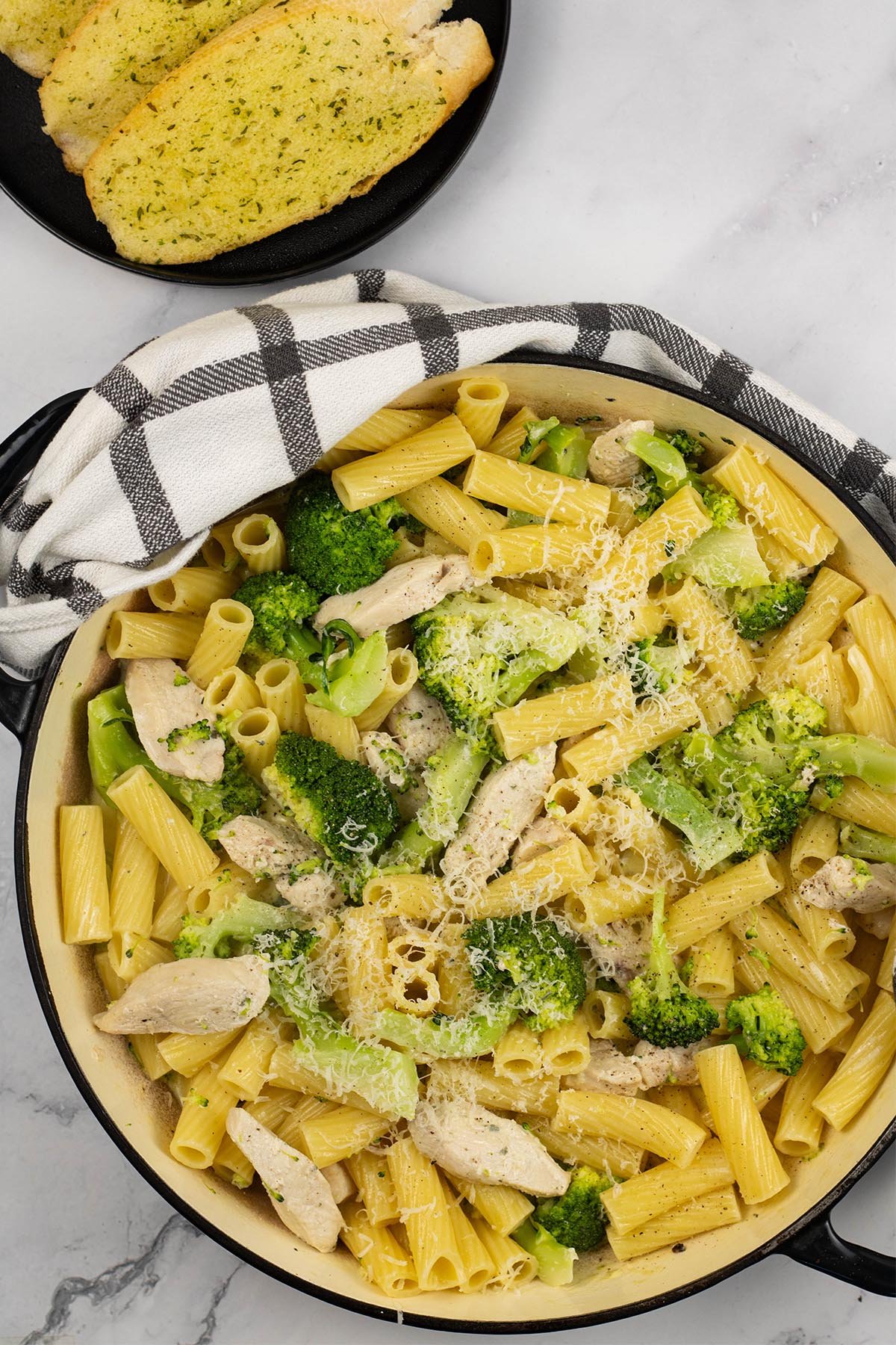 Chicken, broccoli and cheese pasta in large round casserole, with a plate of garlic bread slices in the background.