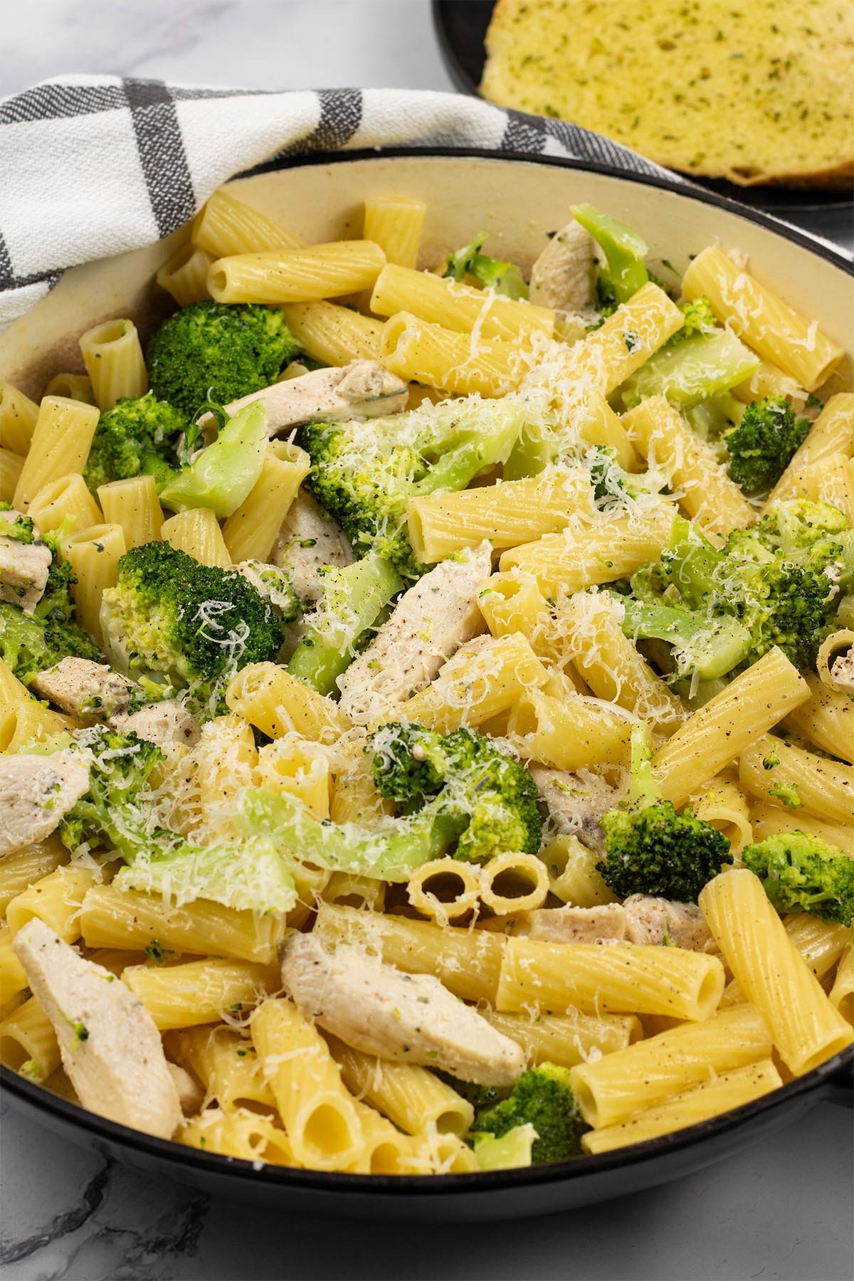 Chicken, broccoli and cheese pasta in large round casserole, with a plate of garlic bread slices in the background.