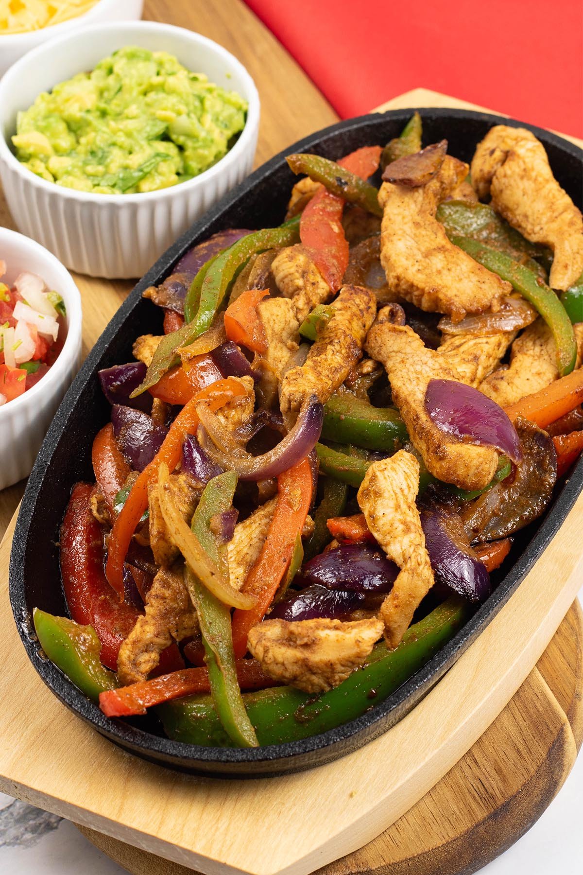 A portion of chicken fajitas in a sizzle platter sitting on a board with small bowls of guacamole, grated cheese and pico de Gallo salsa on the side.