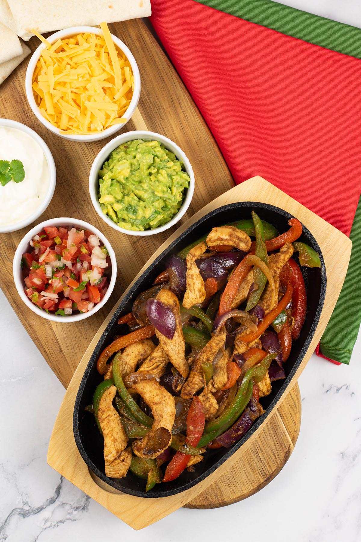 A portion of chicken fajitas in a sizzle platter sitting on a board with small bowls of sour cream, guacamole, grated cheese and pico de gallo salsa, and tortilla wraps on the side.