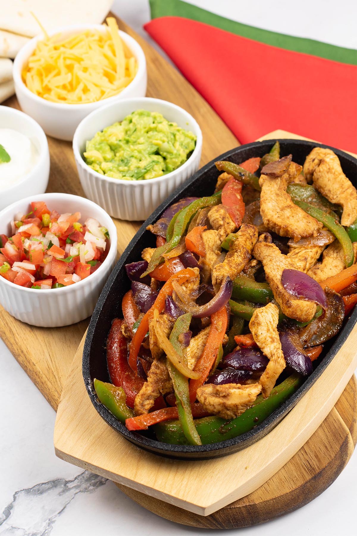 A portion of chicken fajitas in a sizzle platter sitting on a board with small bowls of sour cream, guacamole, grated cheese and pico de gallo salsa, and tortilla wraps on the side.