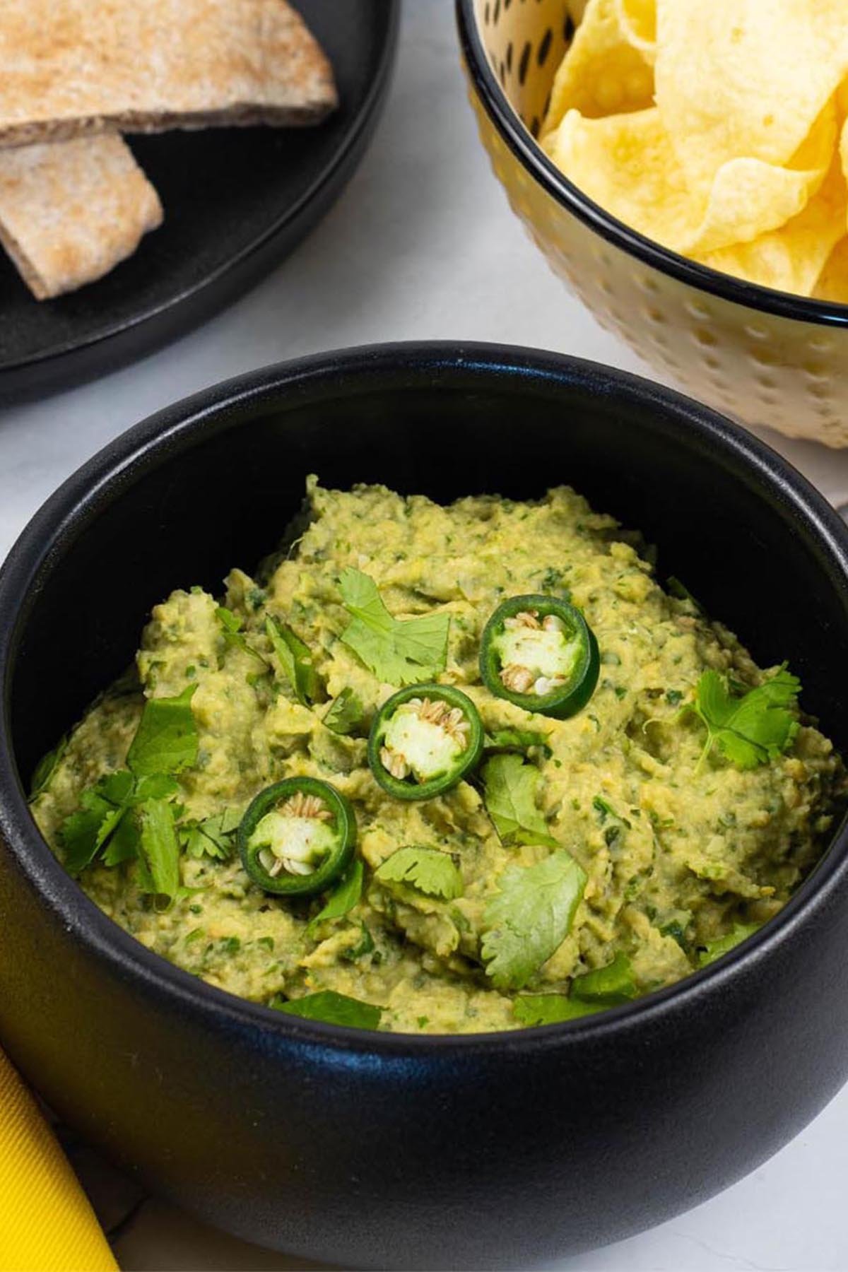 Coriander and green chilli houmous in a black serving bowl garnished with coriander leaves and sliced green chillis.