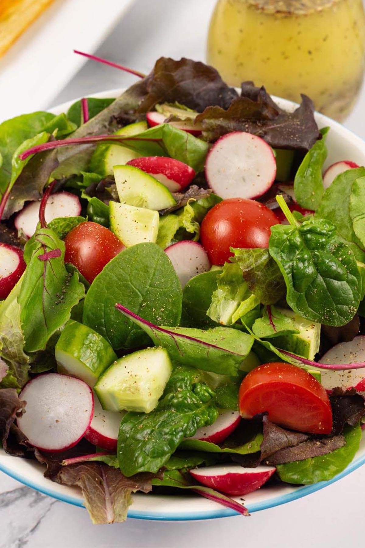 Easy baby leaf salad in a light blue rimmed serving bowl with a jug of zesty Italian-style dressing on the side.