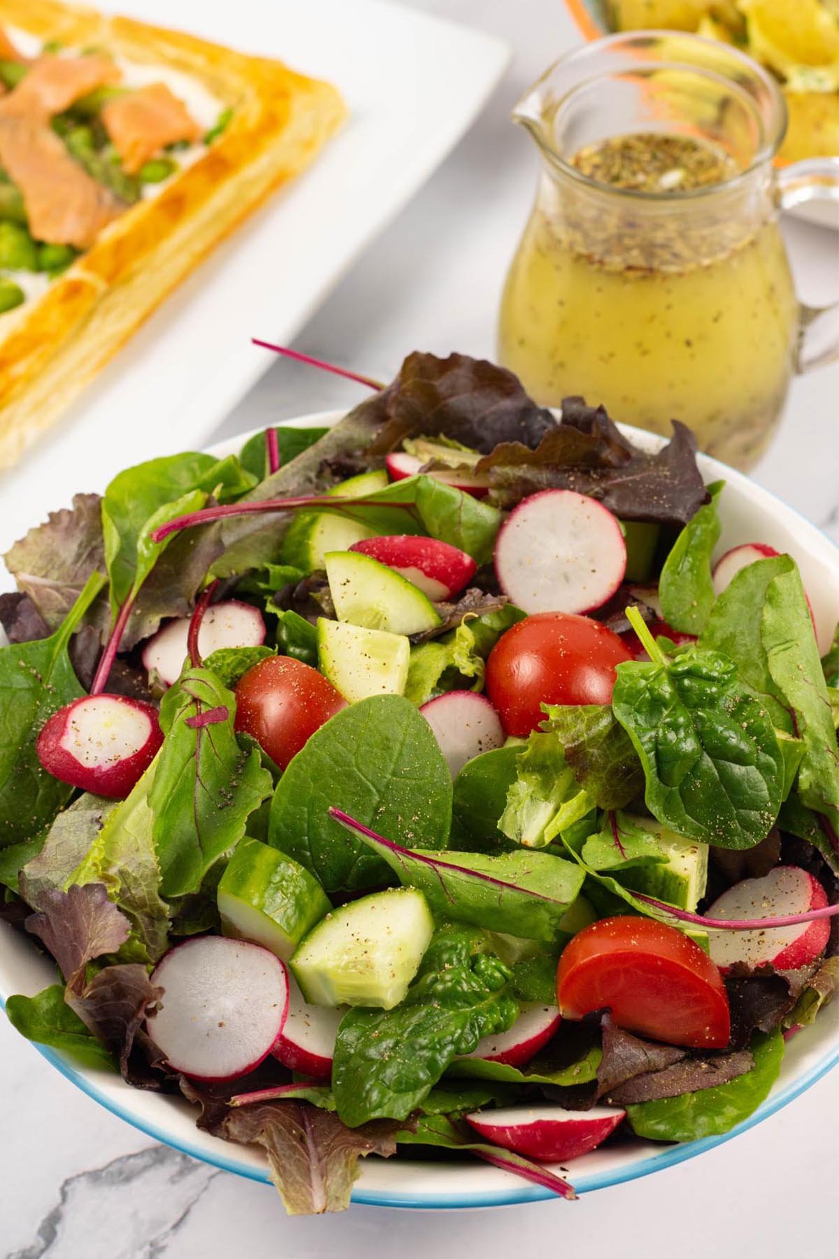 Easy baby leaf salad in a light blue rimmed serving bowl with a jug of zesty Italian-style dressing on the side.