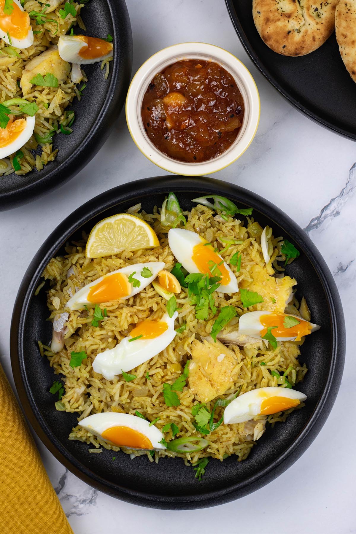Two portions of kedgeree in black bowls with a small dish of mango chutney and plate of naan bread in the background.