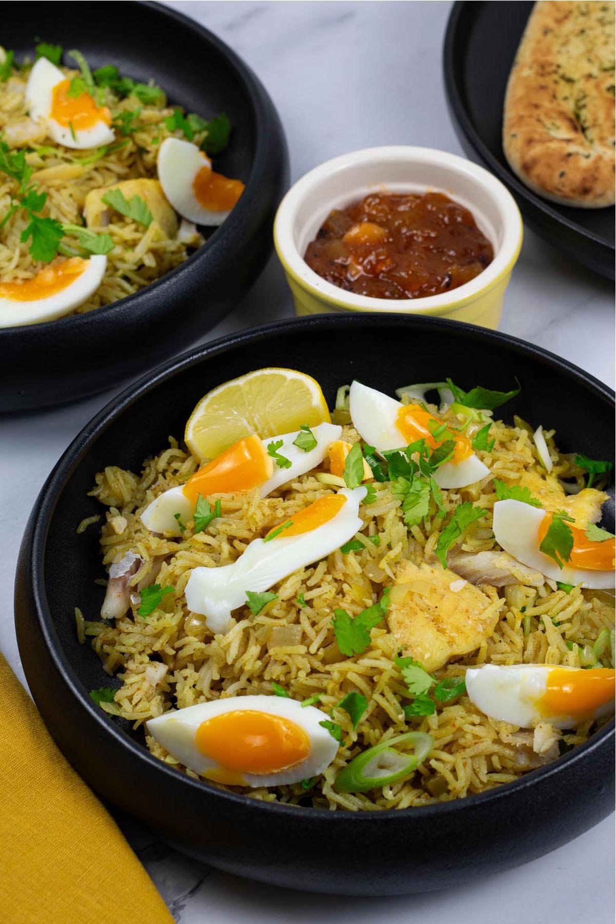 Two portions of kedgeree in black bowls with a small dish of mango chutney and plate of naan bread in the background.