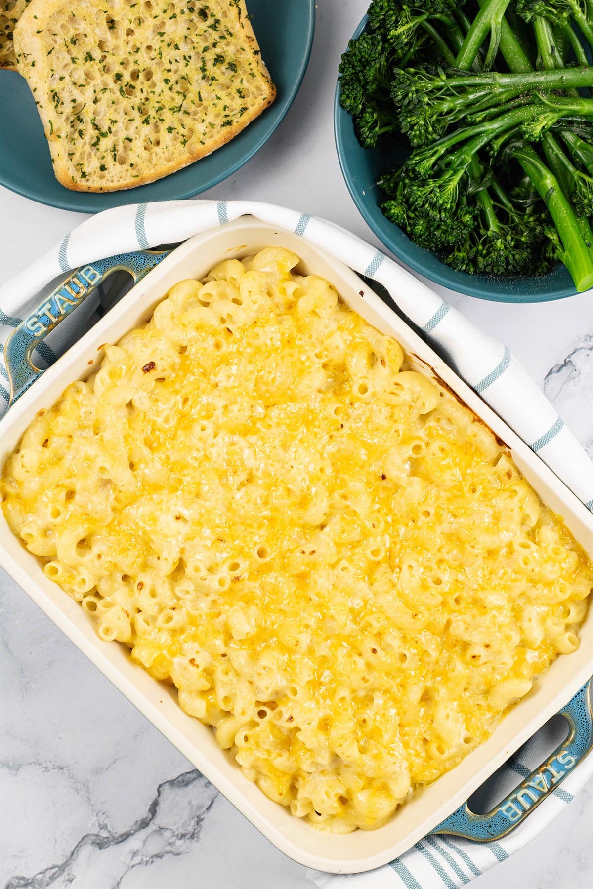 Macaroni cheese in a rectangular Staub dish with a plate of garlic bread and bowl of tenderstem broccoli in the background.