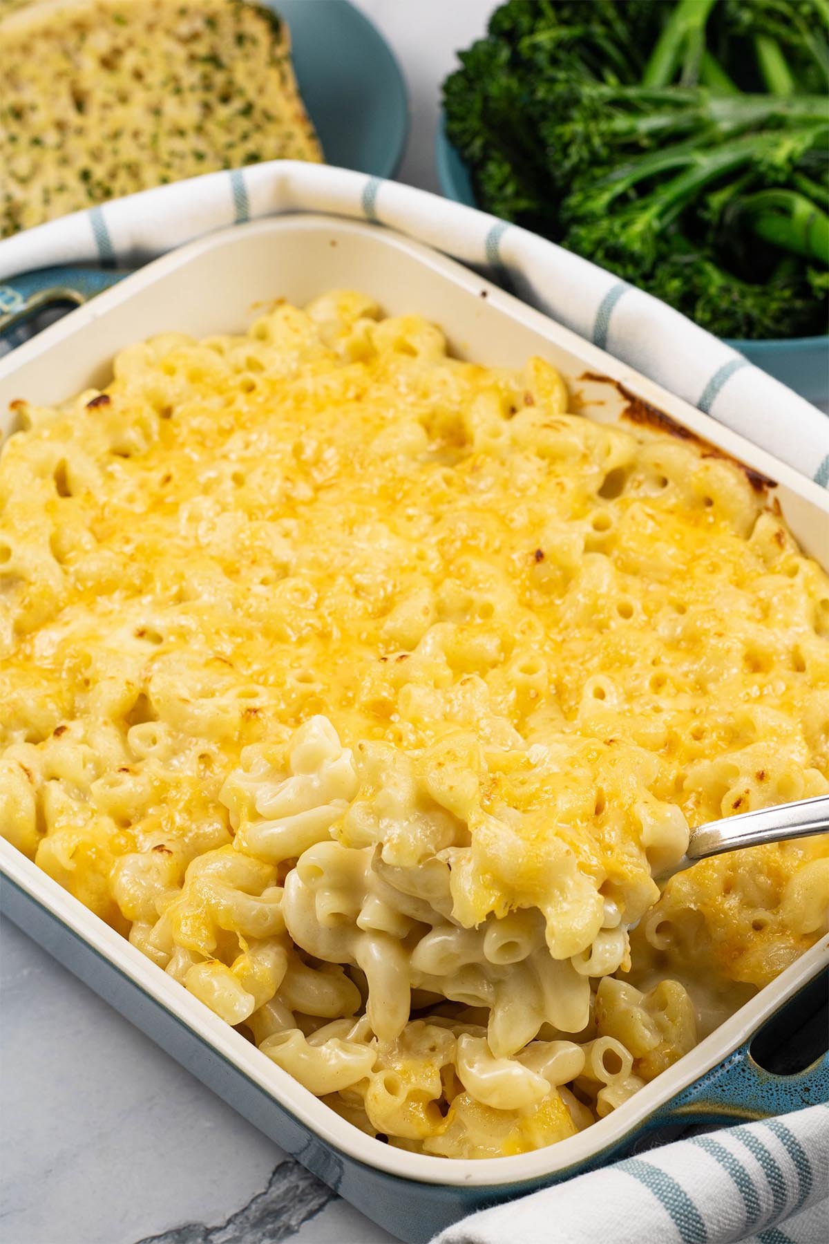 Macaroni cheese in a rectangular Staub dish with a plate of garlic bread and bowl of tenderstem broccoli in the background.