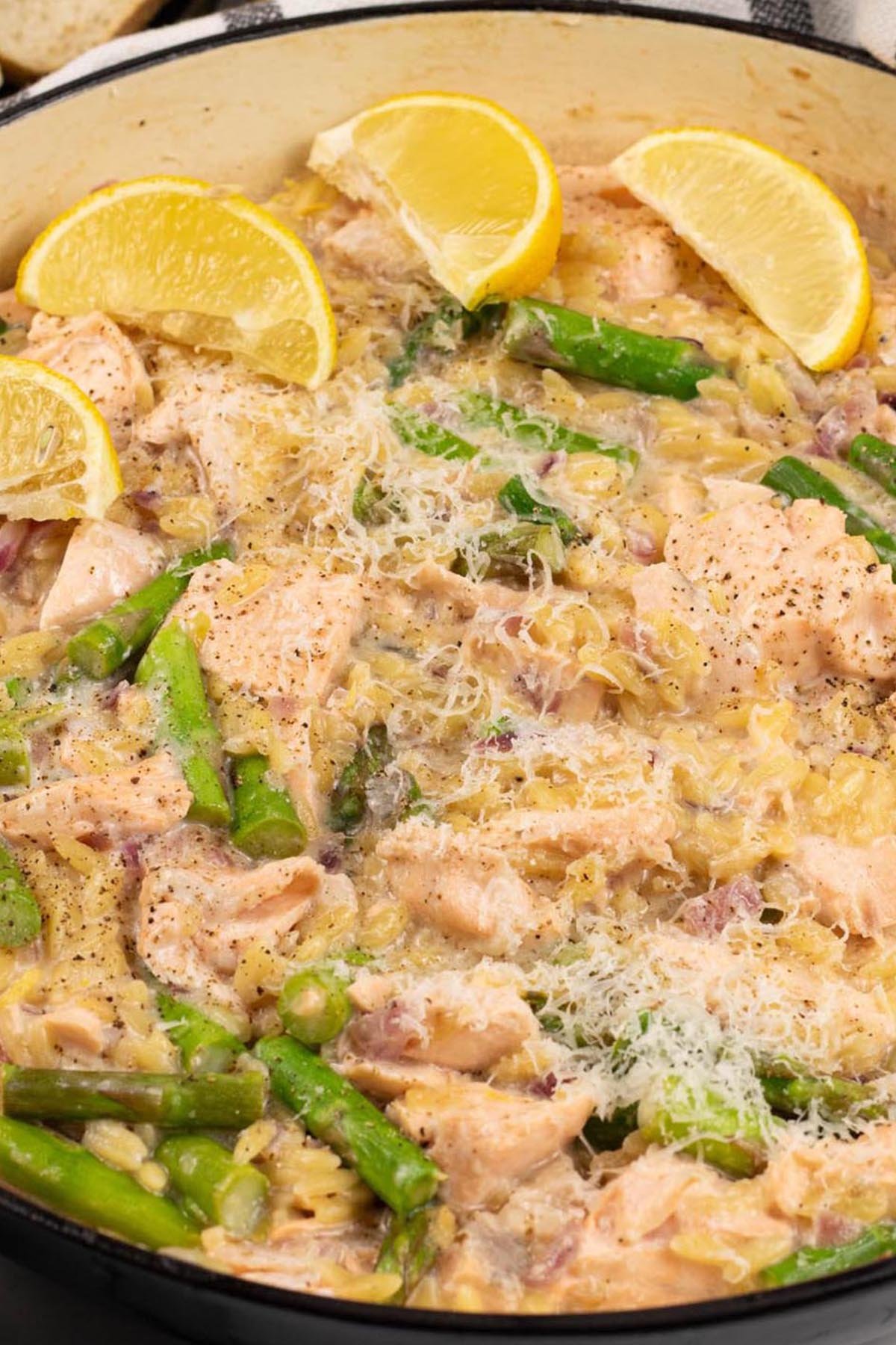 Salmon and asparagus orzo in a large round shallow casserole, garnished with parmesan and lemon wedges.