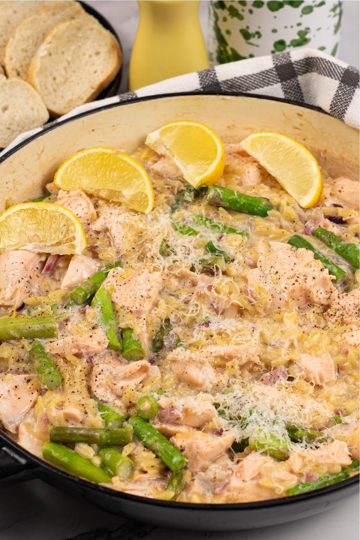 Salmon and asparagus orzo in a large round shallow casserole, garnished with parmesan and lemon wedges.