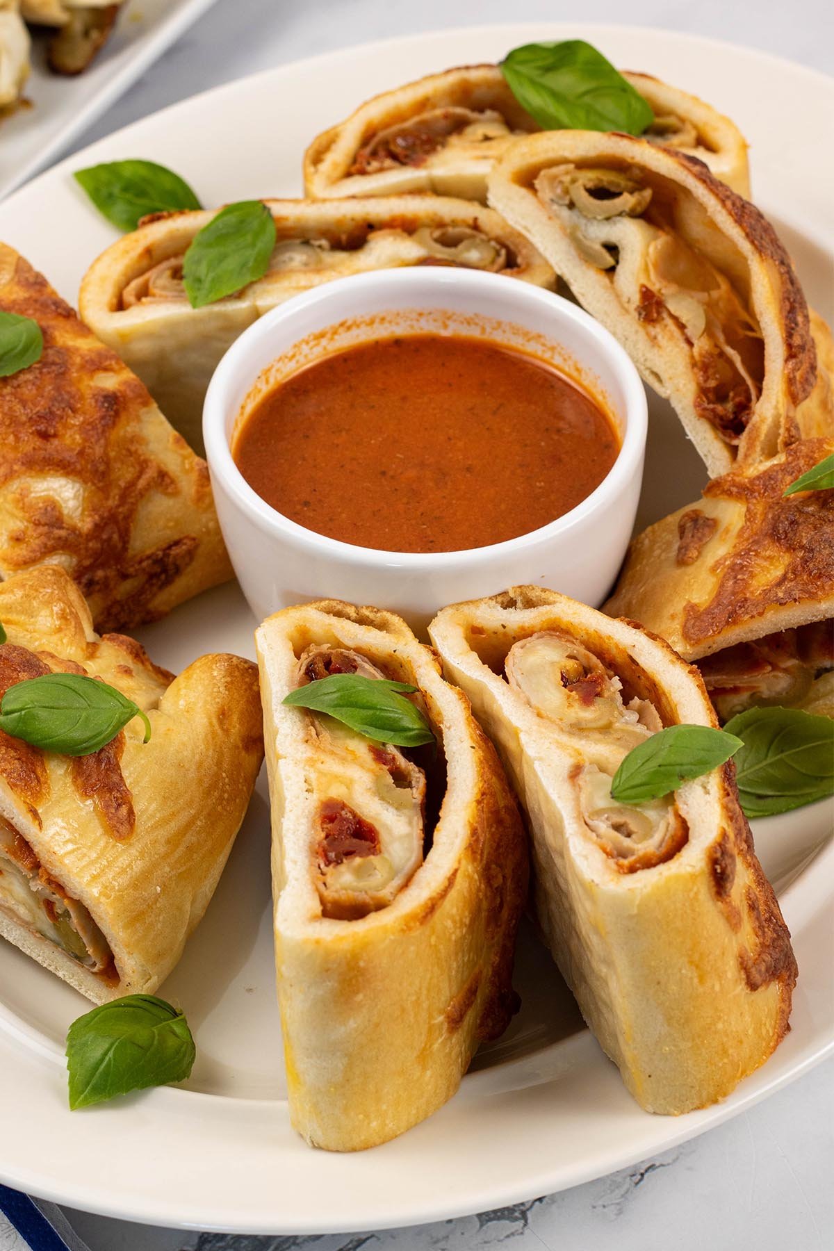 Sliced stromboli on an oval white platter, garnished with basil leaves and served with a dish of tomato pizza sauce.
