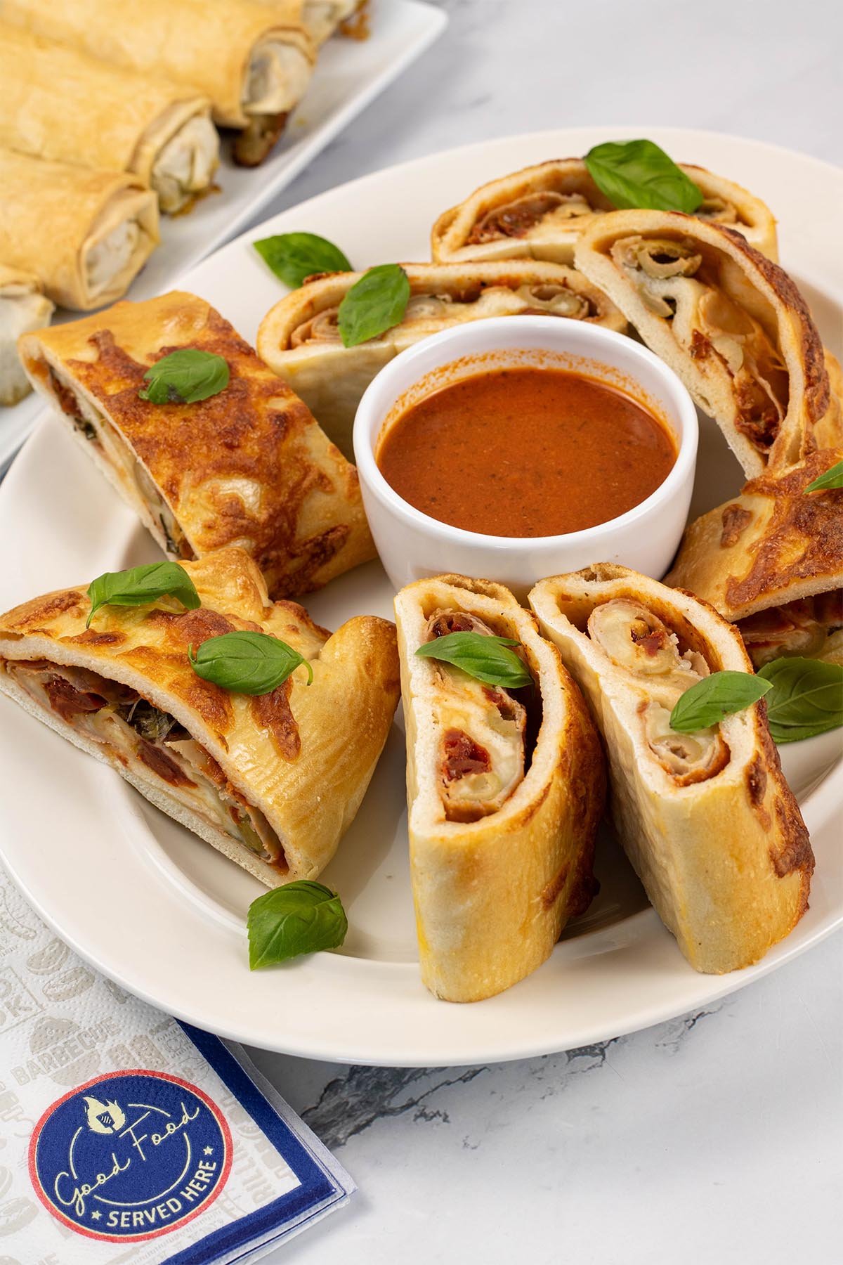 Sliced stromboli on an oval white platter, garnished with basil leaves and served with a dish of tomato pizza sauce.