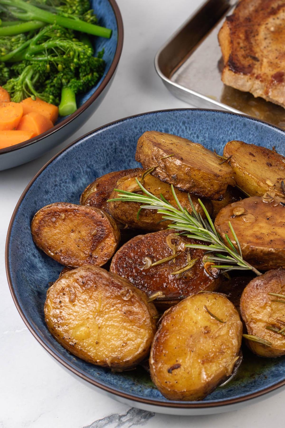 Buttery herbed melting potatoes in a blue patterned serving bowl, with a bowl of carrots and broccoli and tray of pork chops in the background.