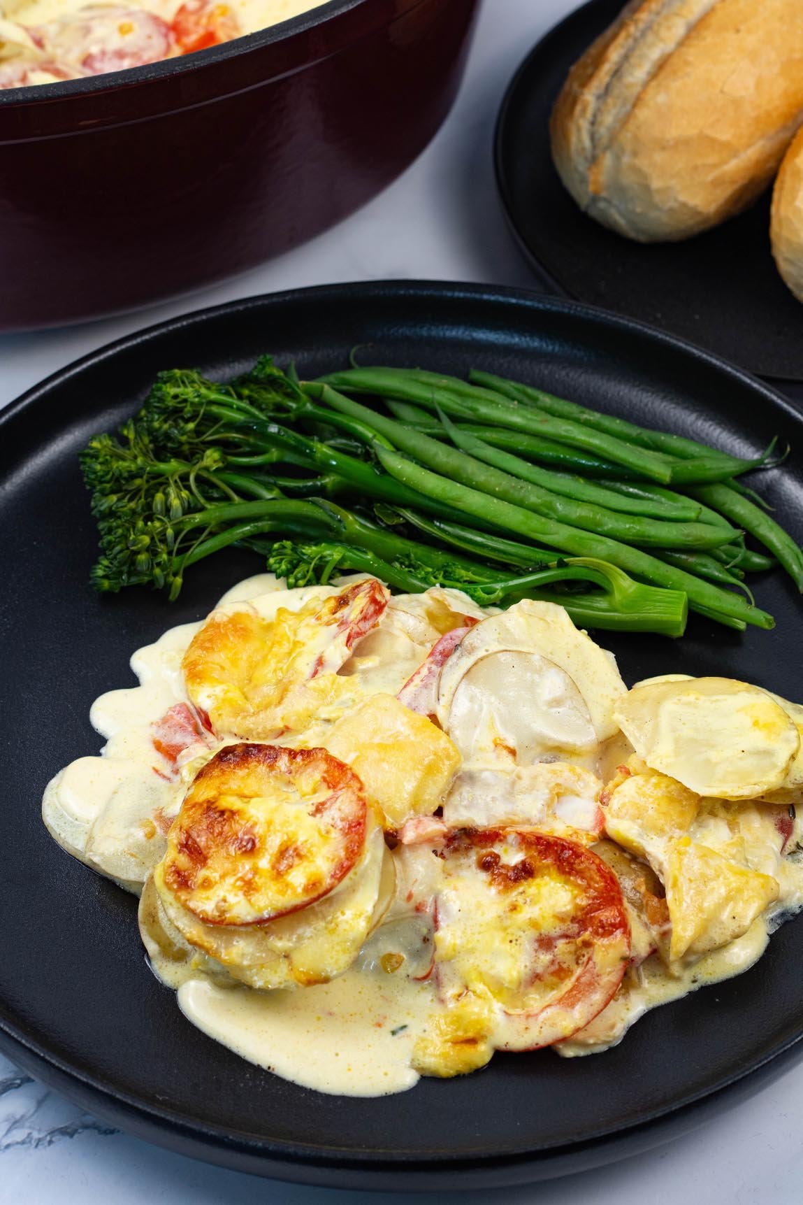 A portion of creamy gratin of smoked haddock, tomatoes and potatoes on a black dinner plate green veg and a plate of crusty rolls in the background.
