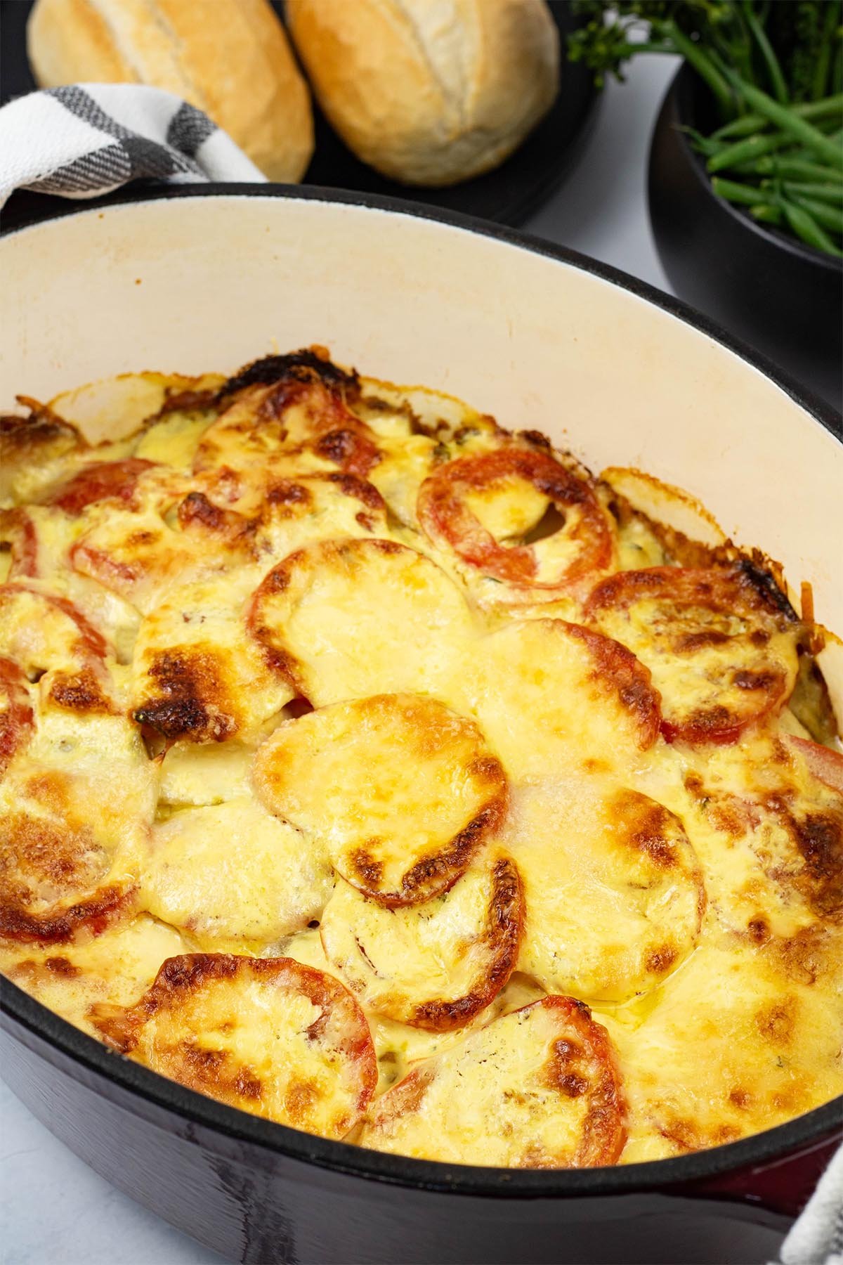Creamy gratin of smoked haddock, tomatoes and potatoes in a deep oval casserole dish with crusty rolls and green veg in the background.