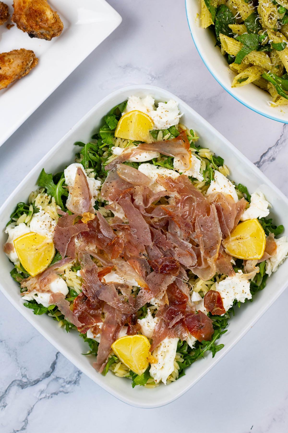 Crispy prosciutto and orzo salad in a white square serving dish with a bowl of pesto, peas and spinach pasta salad, and platter of cajun wings in the background.
