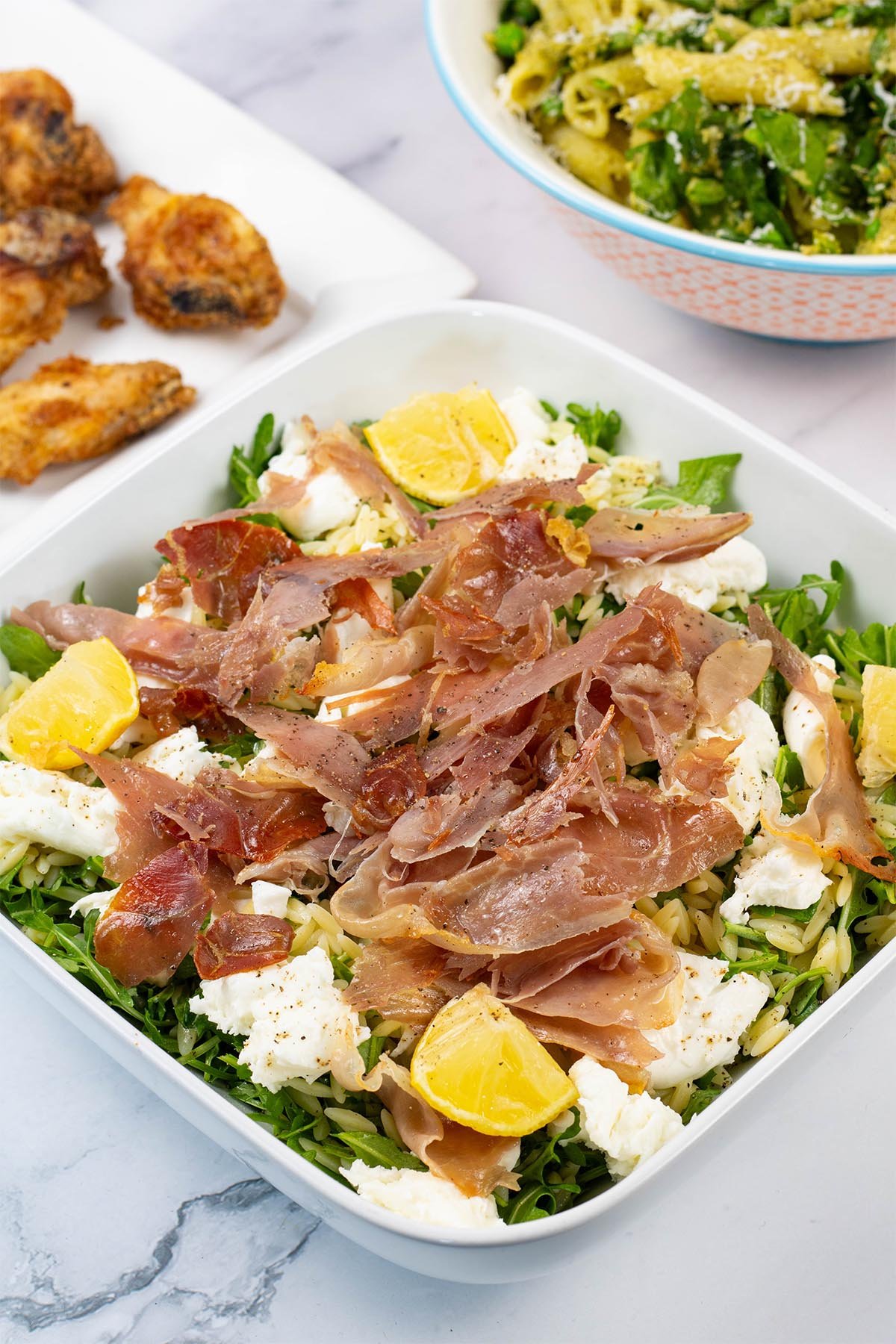 Crispy prosciutto and orzo salad in a white square serving dish with a bowl of pesto, peas and spinach pasta salad, and platter of cajun wings in the background.