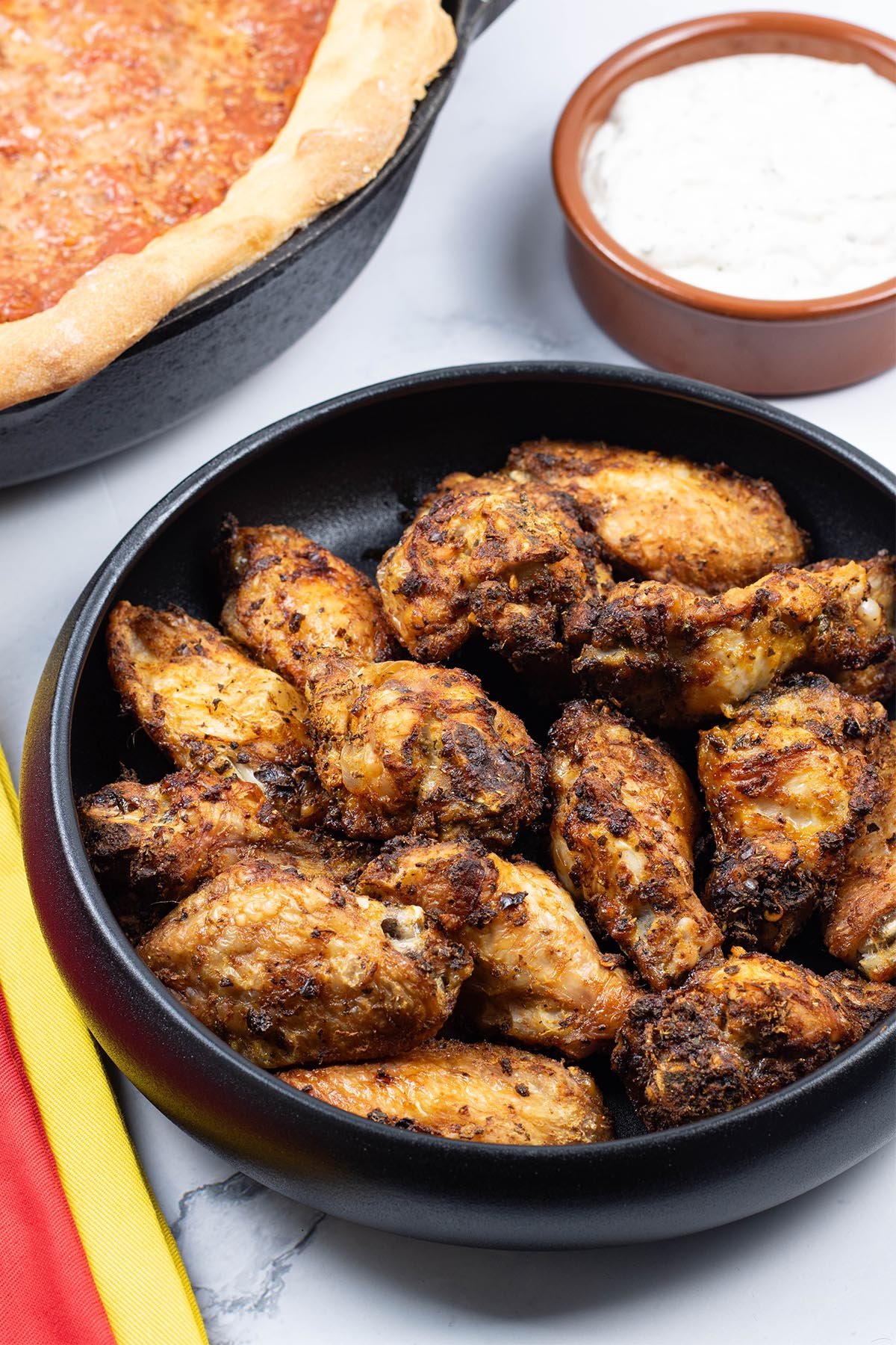 Easy air fryer Spanish chicken wings in a black serving bowl with a Chicago deep pan pizza and bowl of garlic and herb dip in the background.