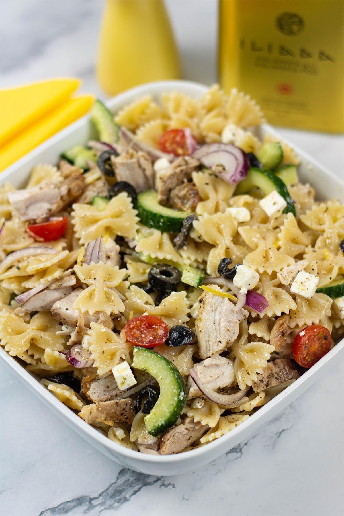 Greek chicken pasta salad in a square white serving dish with a yellow pepper grinder and tin of Greek extra virgin olive oil in the background.