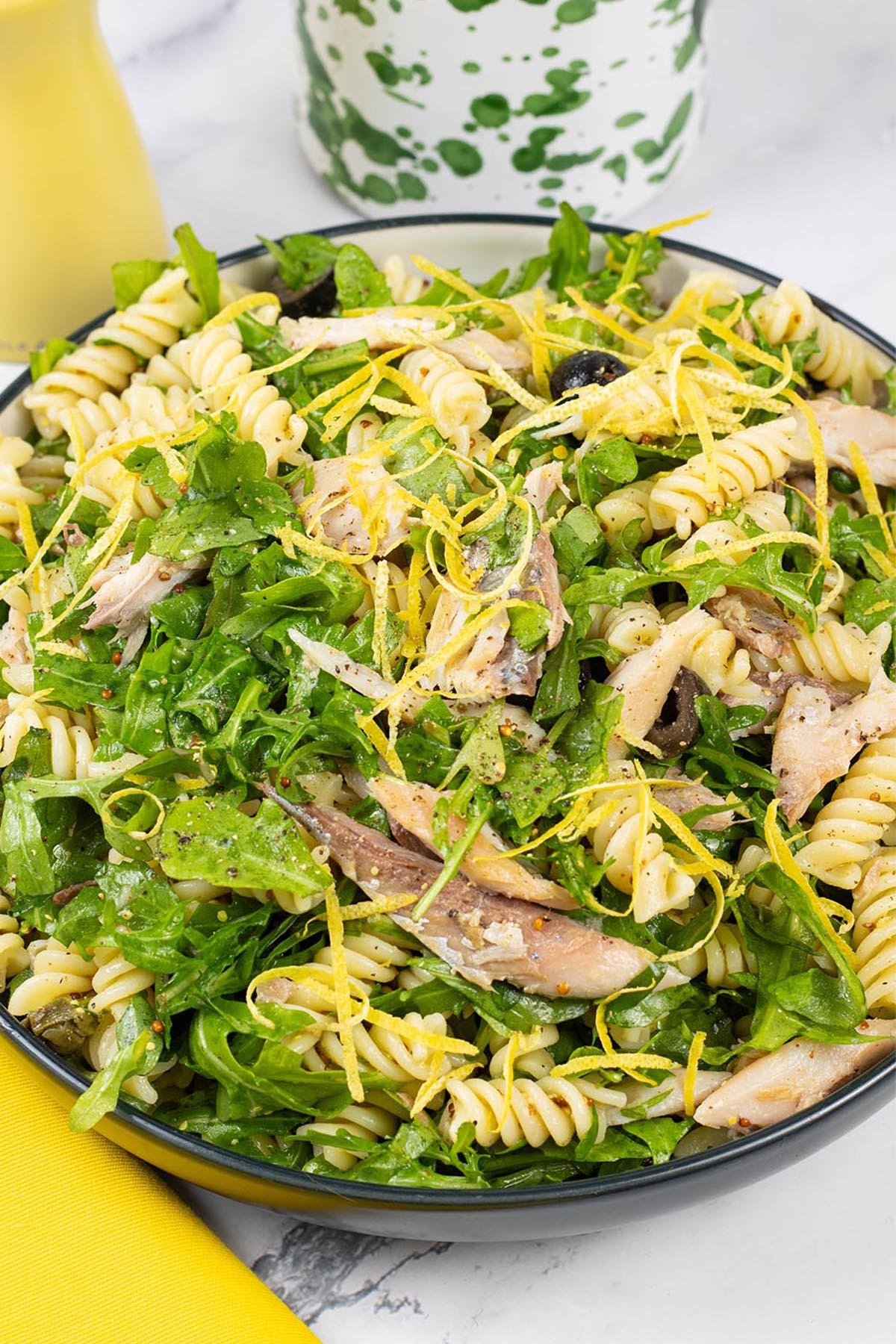 Hot smoked mackerel and rocket pasta salad in a black and cream serving bowl with a yellow pepper grinder and green and white oil bottle in the background.