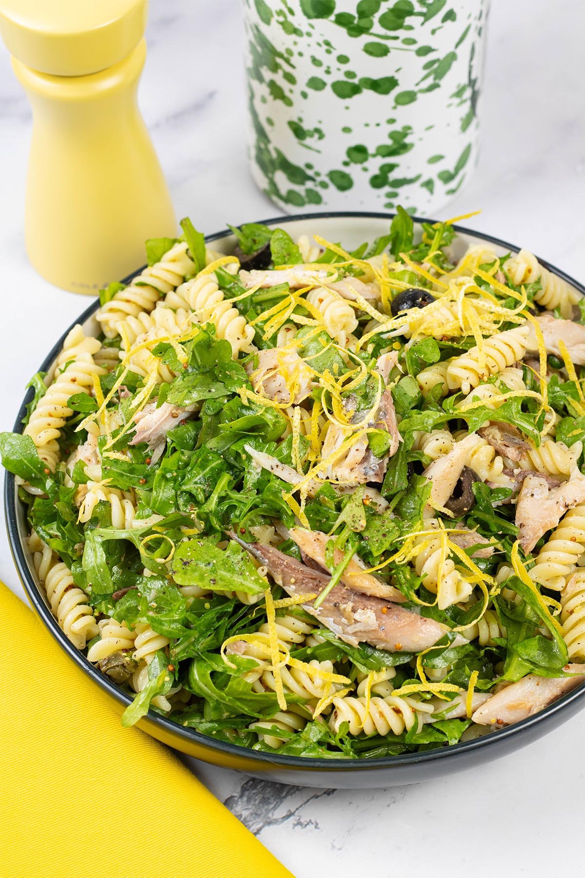 Hot smoked mackerel and rocket pasta salad in a black and cream serving bowl with a yellow pepper grinder and green and white oil bottle in the background.