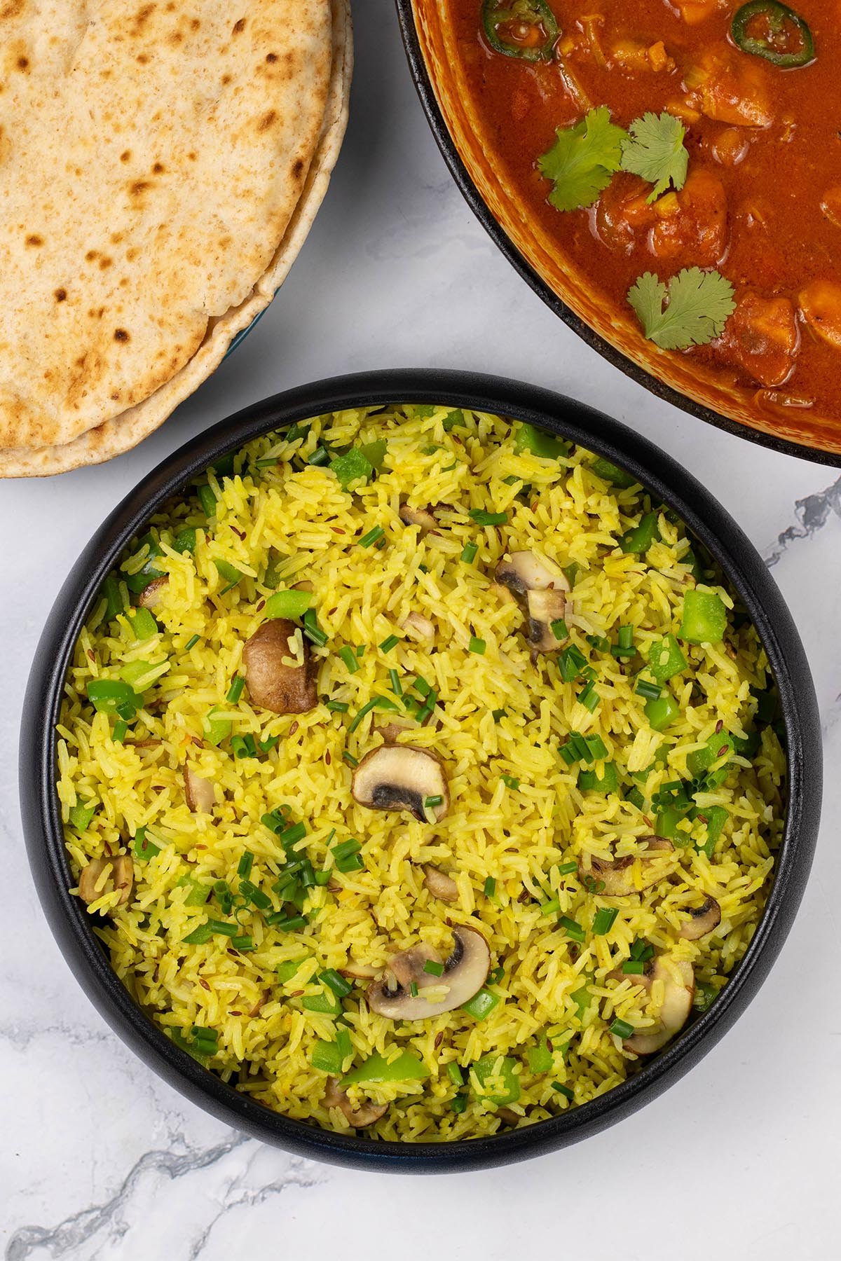 Mushroom rice in a black serving bowl with a plate of chapatis and dish of speedy gunpowder chicken in the background.