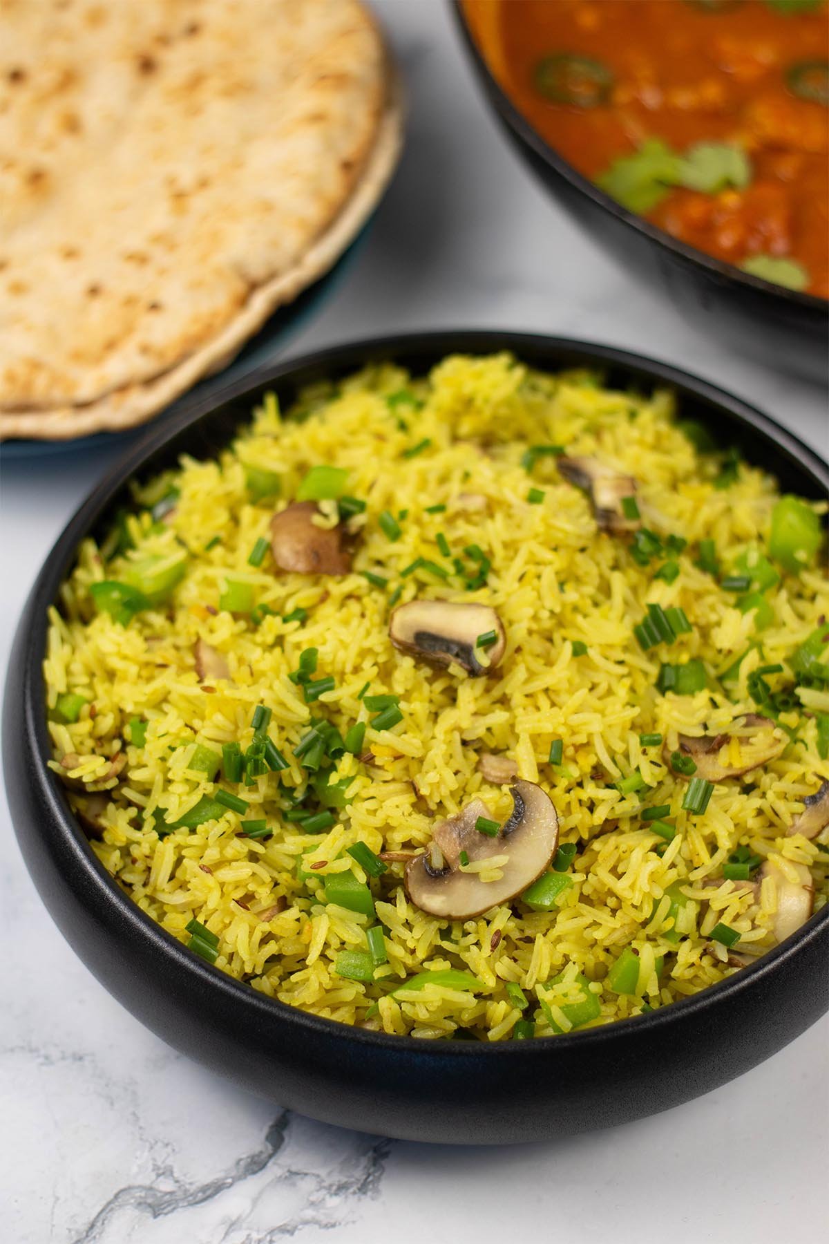 Mushroom rice in a black serving bowl with a plate of chapatis and dish of speedy gunpowder chicken in the background.
