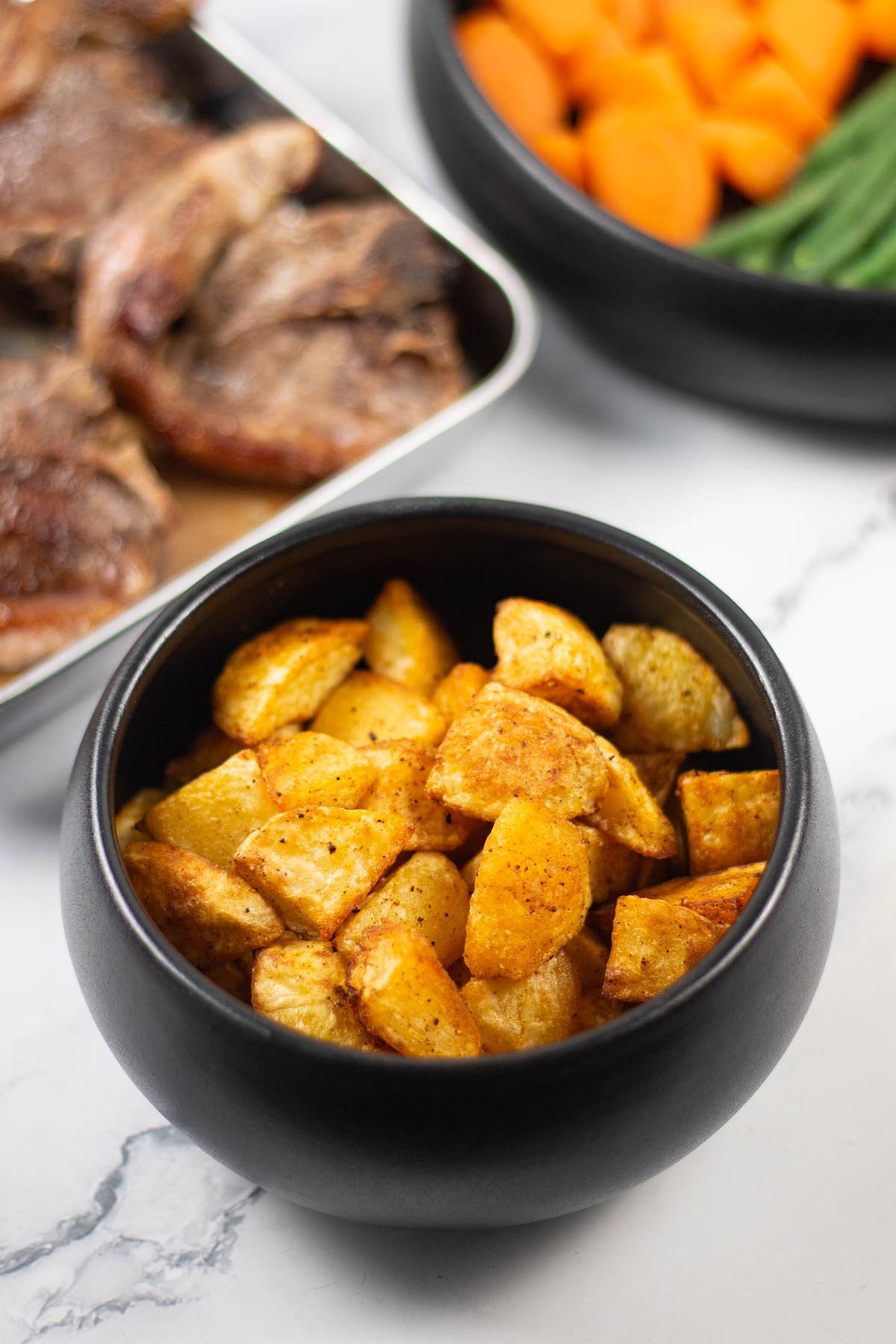 Paprika parmentier potatoes in a black serving bowl with pan-fried lamb chops and a bowl of carrots and green beans in the background.