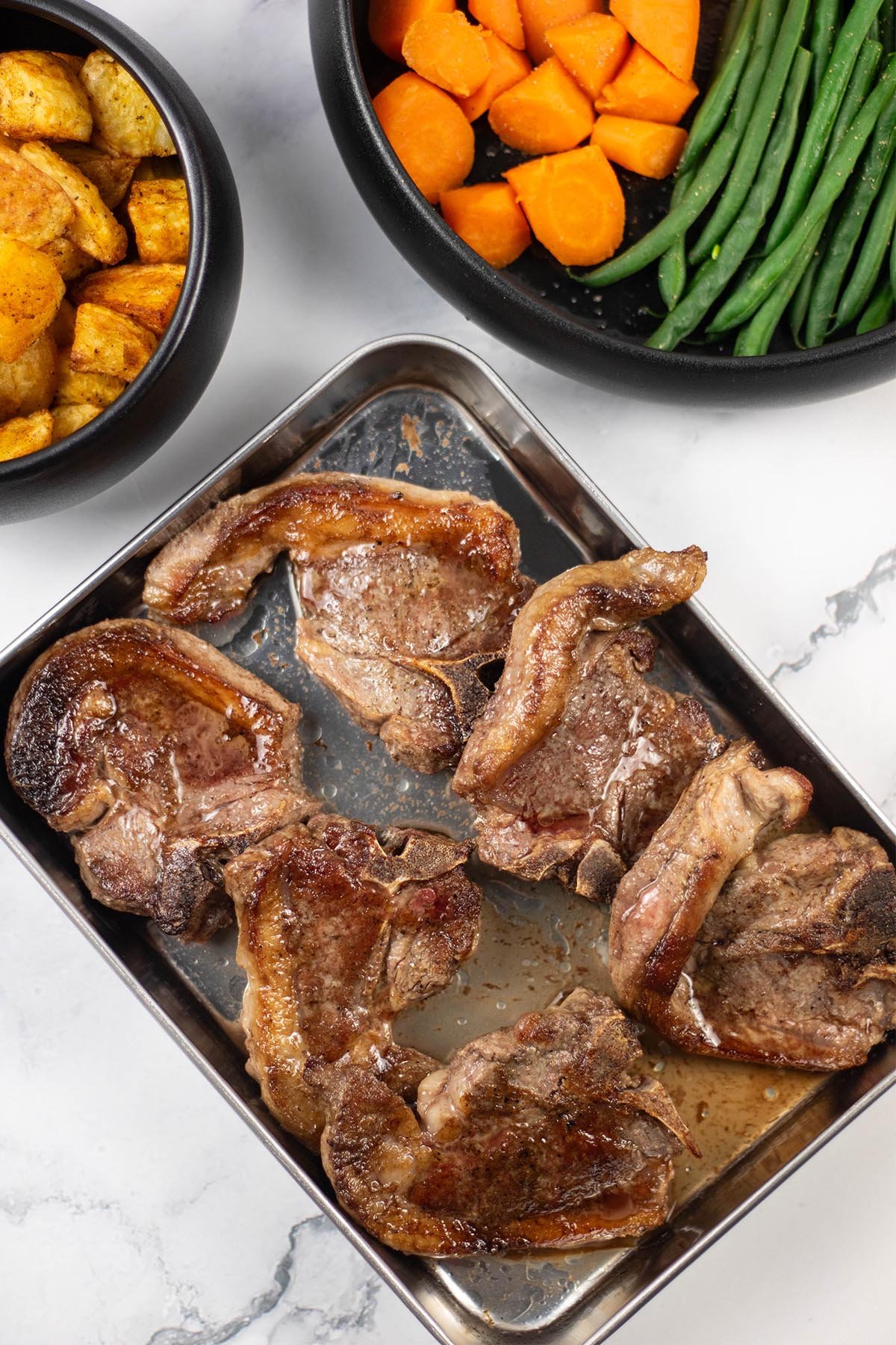 6 perfect pan-fried lamb chops resting on a tray with bowls of parmentier potatoes, carrots and green beans in the background.
