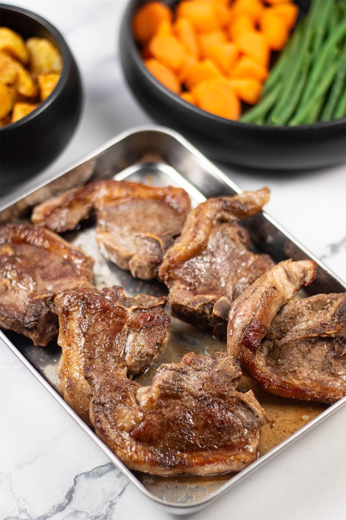 6 perfect pan-fried lamb chops resting on a tray with bowls of parmentier potatoes, carrots and green beans in the background.