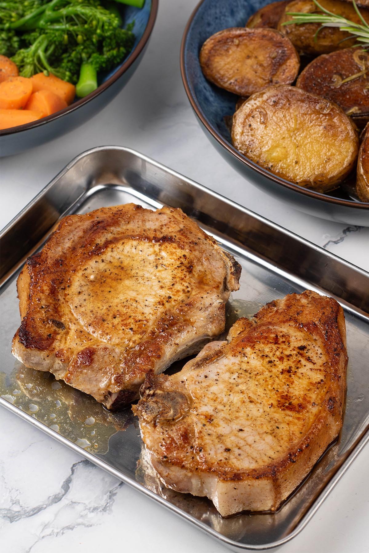 2 perfect pan-fried pork chops resting on a tray with bowls of melting potatoes, carrots and broccoli in the background.